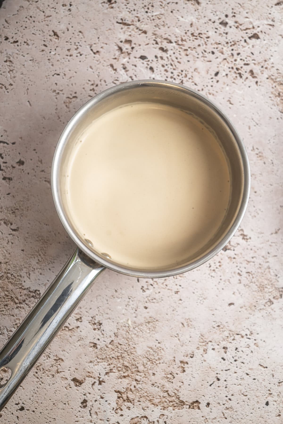 A stainless steel saucepan filled with coffee cream, placed on a textured pinkish-beige surface.