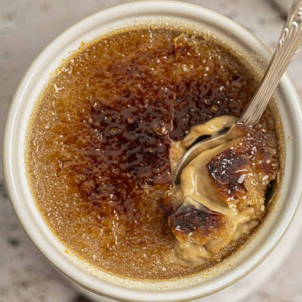 A close-up of a spoon breaking into the caramelized sugar top of a creamy coffee crème brûlée in a white ramekin.