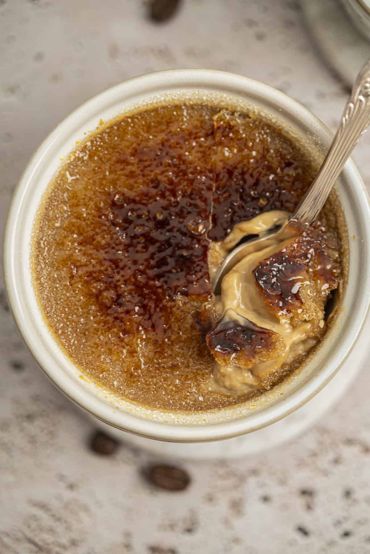 A close-up of a coffee crème brûlée in a white ramekin, with a caramelized sugar crust being cracked by a spoon to reveal creamy custard underneath. Coffee beans are scattered on the textured surface in the background.