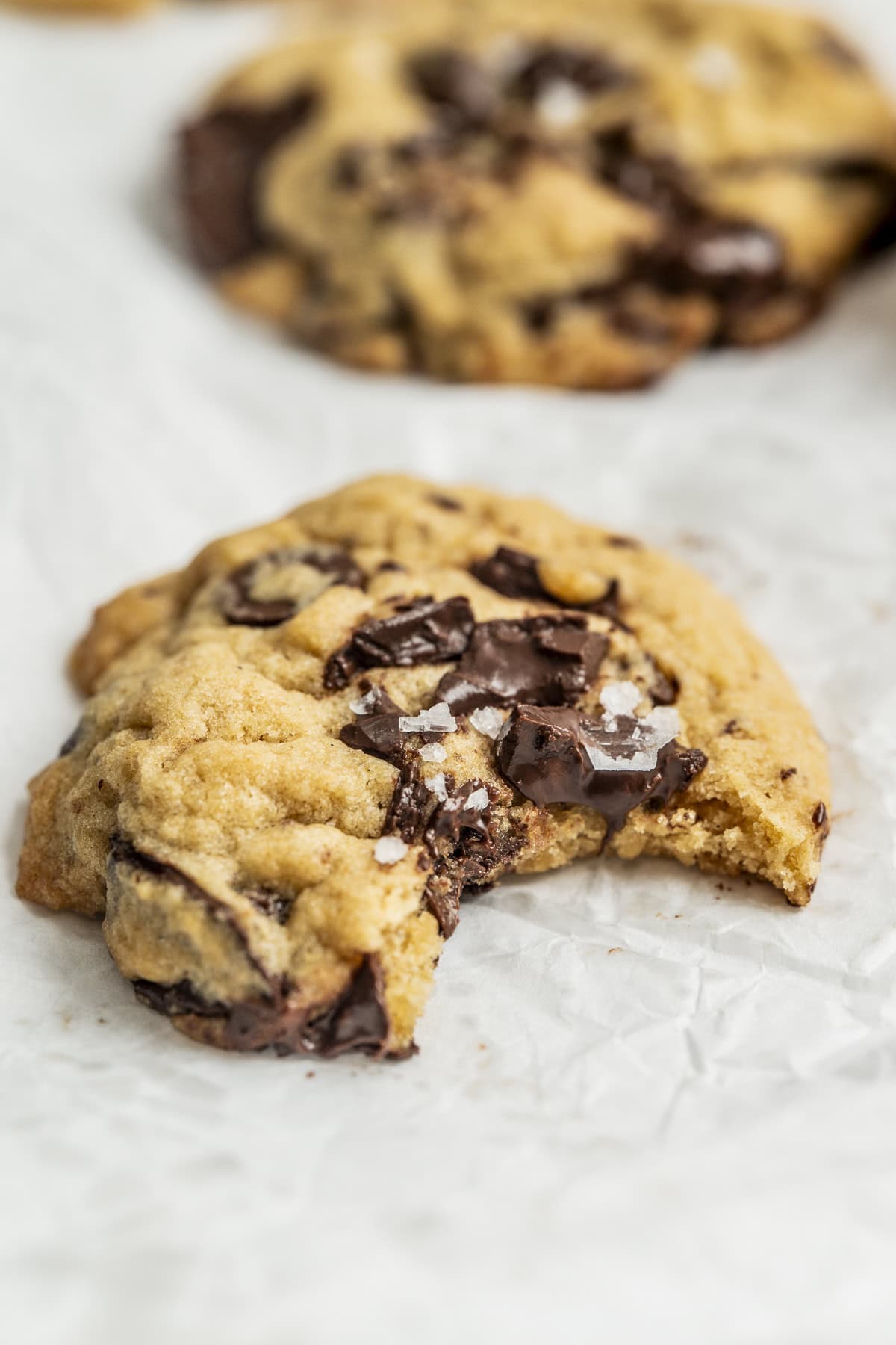 A thick chocolate chip cookie with large melted chocolate chunks and a sprinkle of sea salt sits on crinkled parchment paper, with a bite taken out of it.