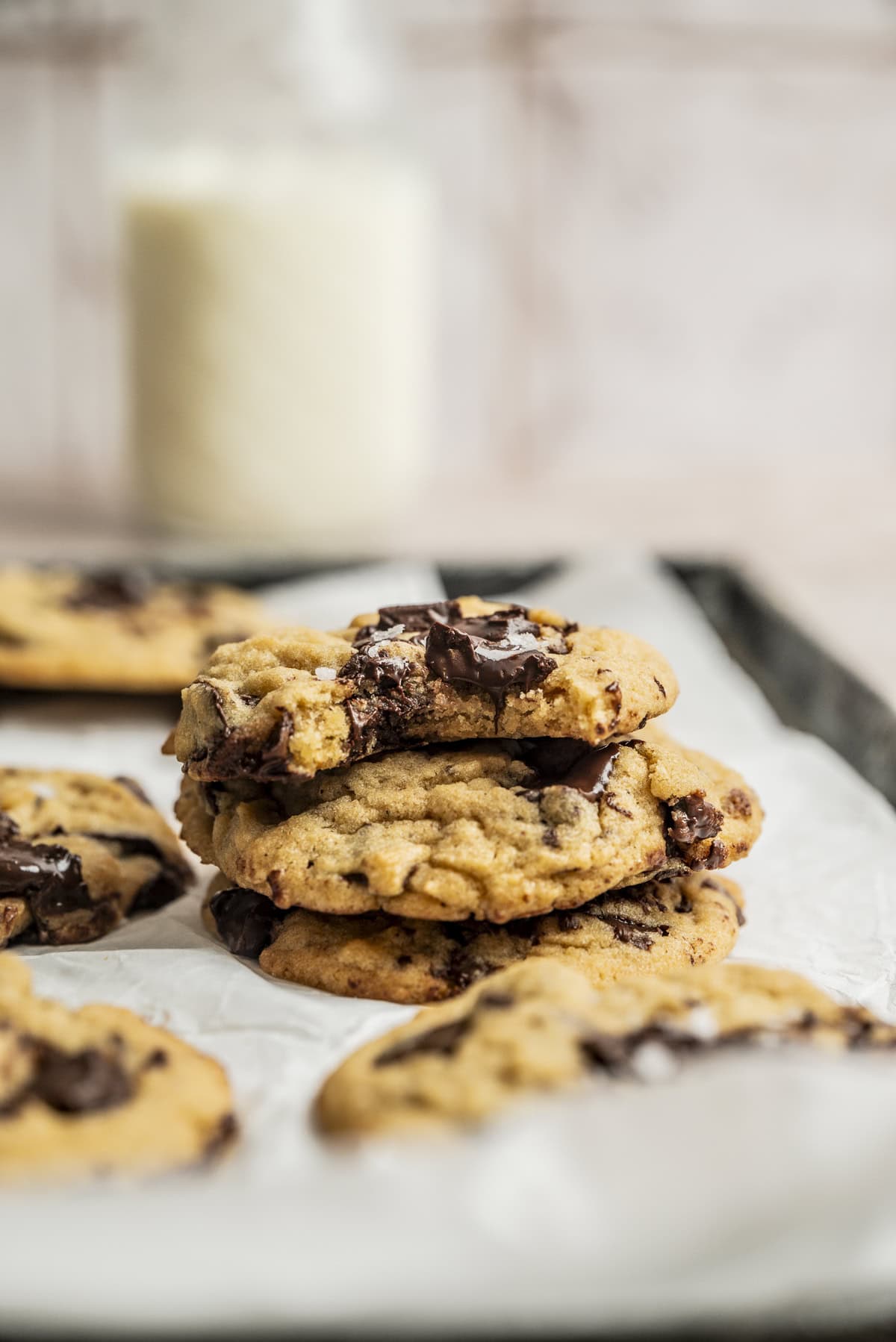 A stack of chocolate chip cookies sits on a baking tray lined with parchment paper, with more cookies in the background and a blurred glass of milk behind them.