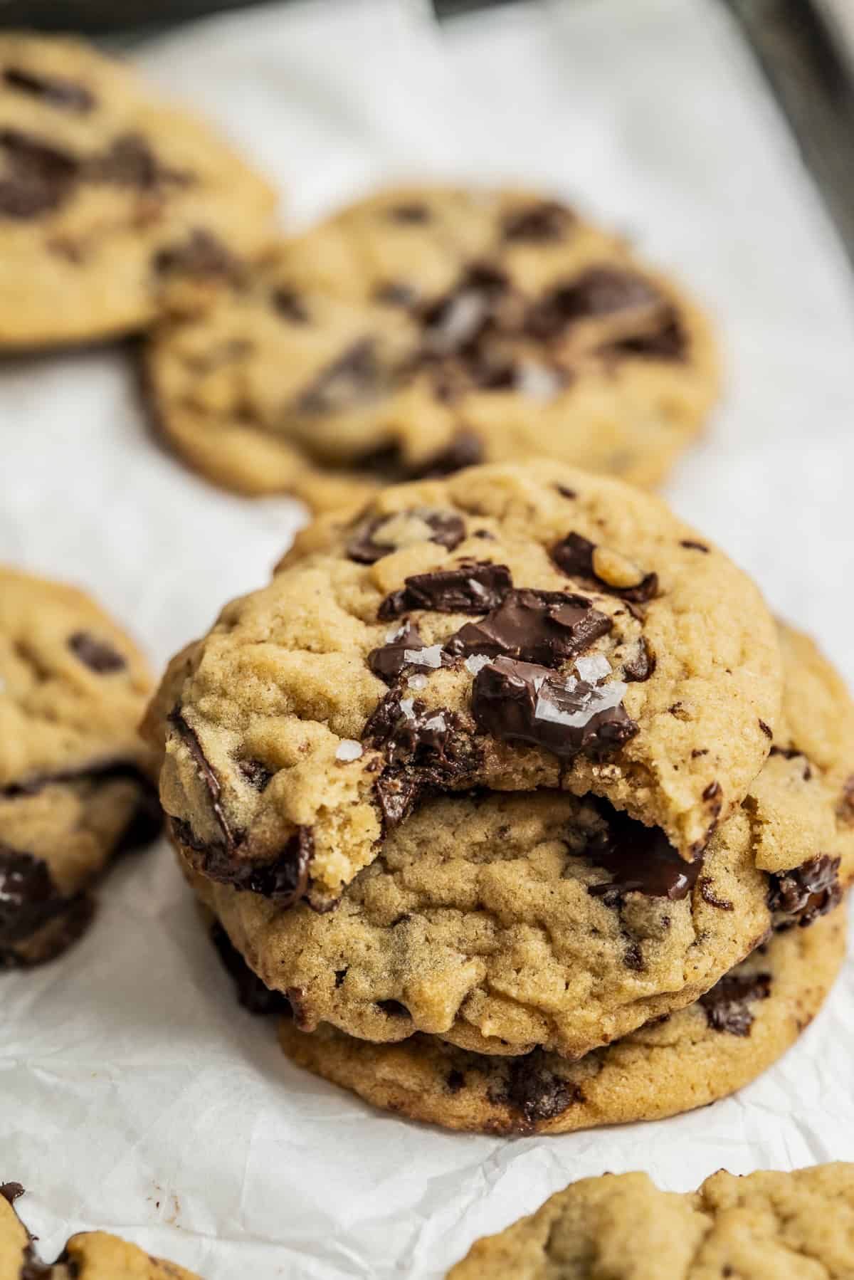 A stack of chocolate chip cookies sits on crumpled parchment paper, with one cookie on top showing a bite taken out, revealing gooey chocolate chunks inside. Other cookies are visible in the background.