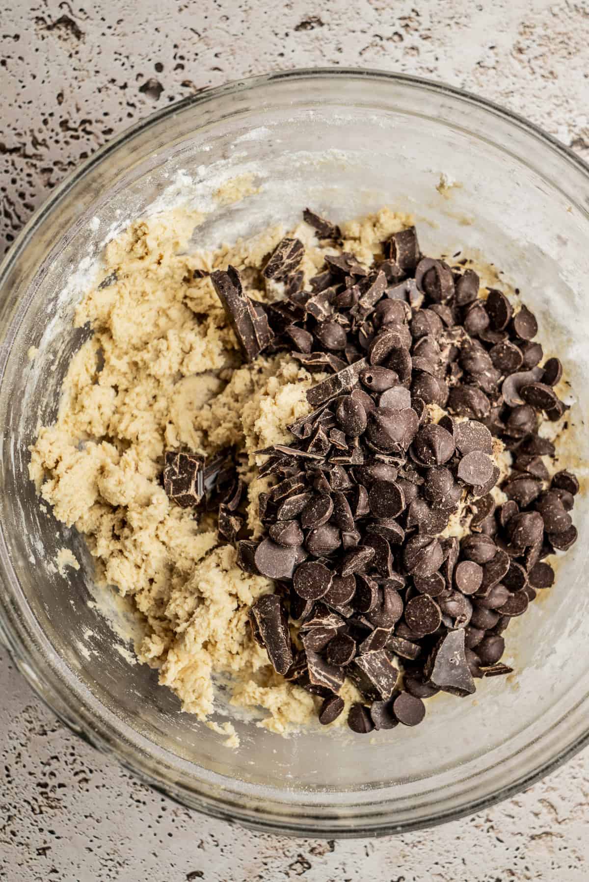 A glass bowl filled with cookie dough, topped with chocolate chips and chocolate chunks, sitting on a textured countertop.