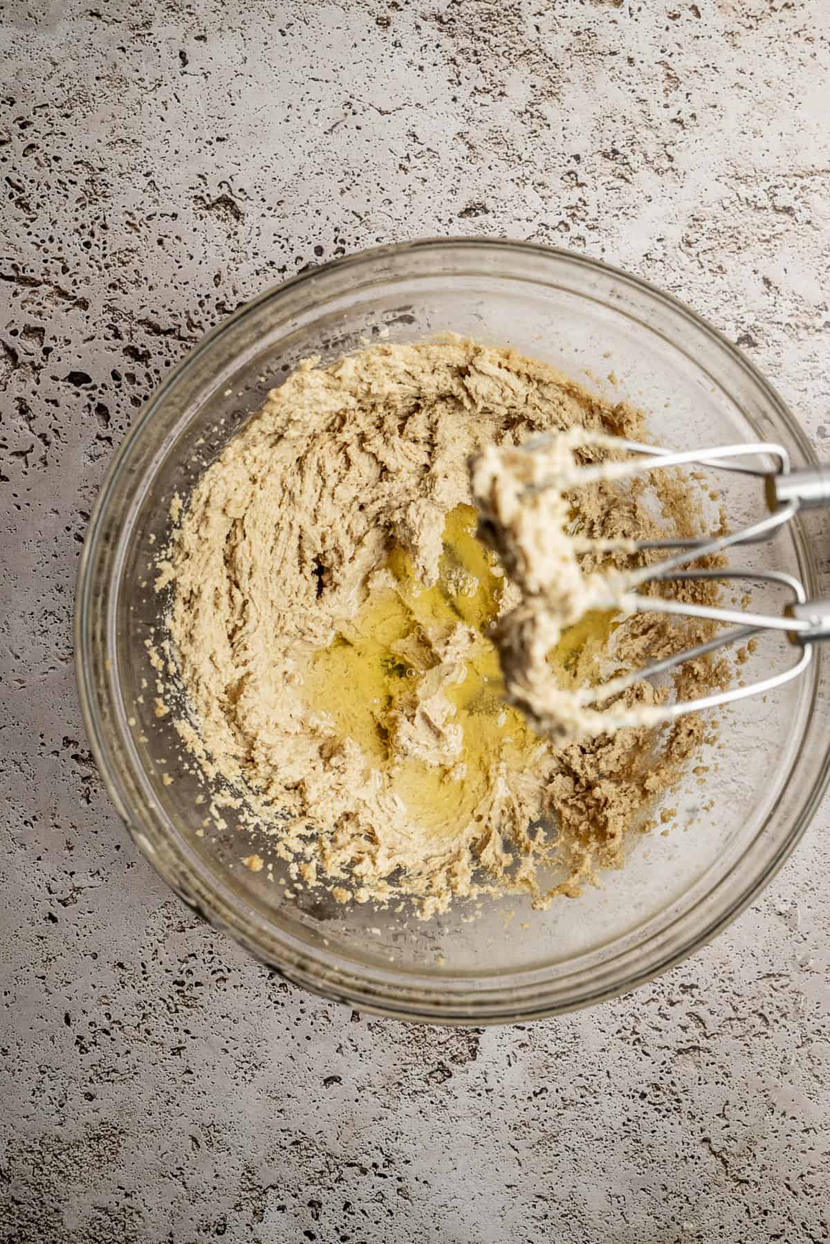 A glass bowl filled with creamed butter and sugar mixture, with an electric hand mixer resting inside, on a speckled countertop.