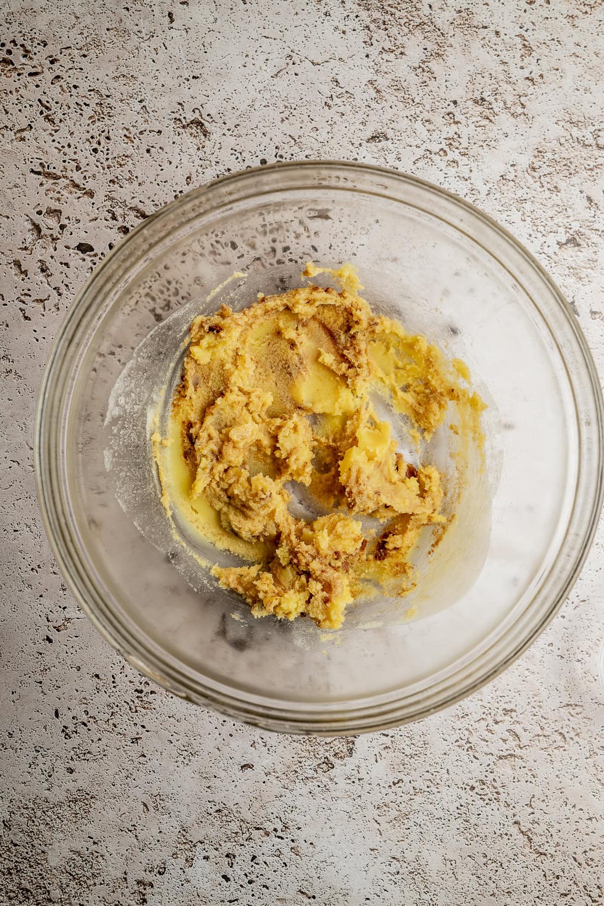 A glass bowl containing creamed butter and brown sugar mixture sits on a light textured surface, viewed from above.