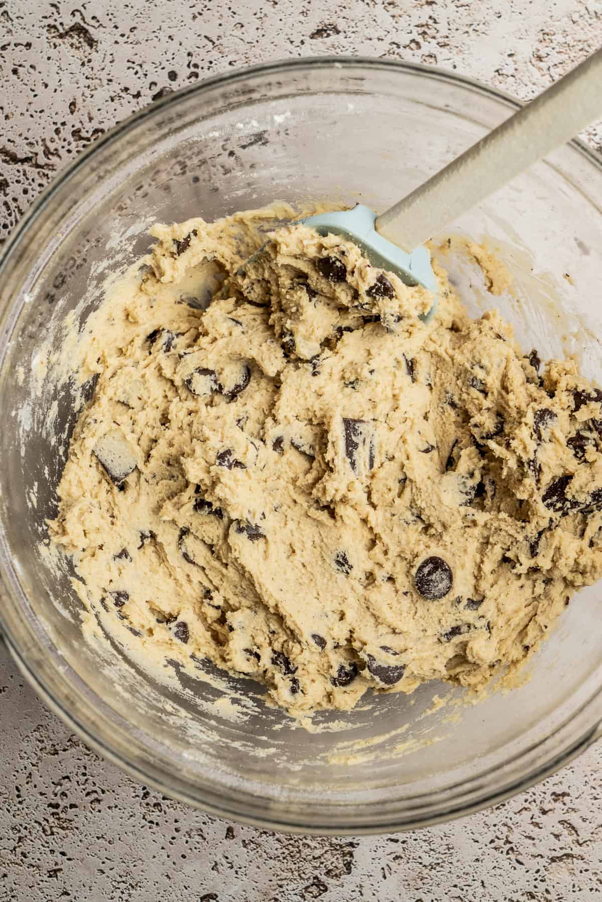 A glass bowl filled with chocolate chip cookie dough, mixed with chocolate chunks, sits on a textured surface. A spatula rests in the dough, ready for scooping or further mixing.