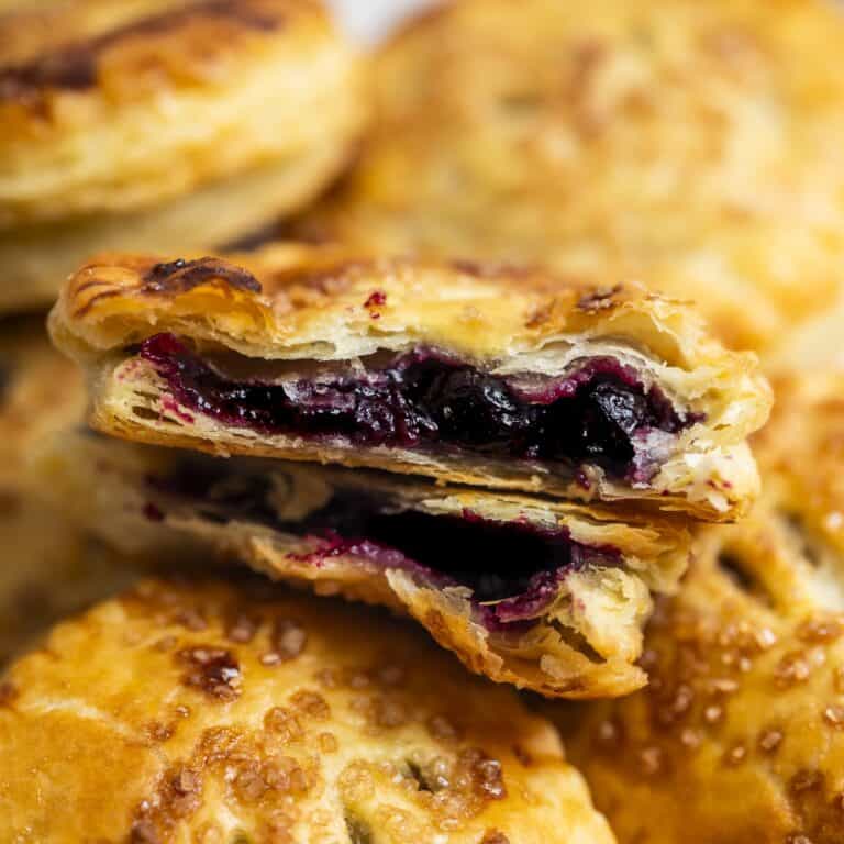 A close-up of a golden, flaky Blackberry Hand Pie filled with dark berry filling, broken open to reveal the juicy fruit inside. Similar pastries are blurred in the background.