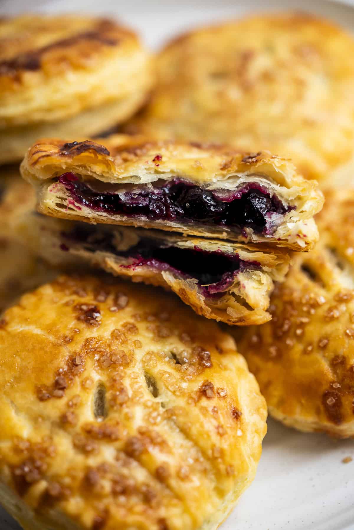 A close-up of golden, flaky blueberry hand pies with pie crust filled with a dark berry filling, with one pie broken open to reveal the juicy filling inside. The pies are arranged on a white plate.