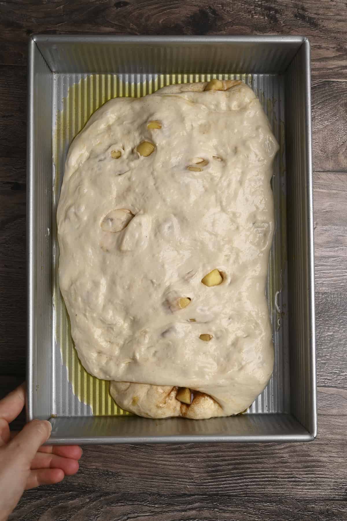 A hand presses raw, uneven pumpkin focaccia dough into a rectangular metal baking pan coated with olive oil, on a dark wooden surface.