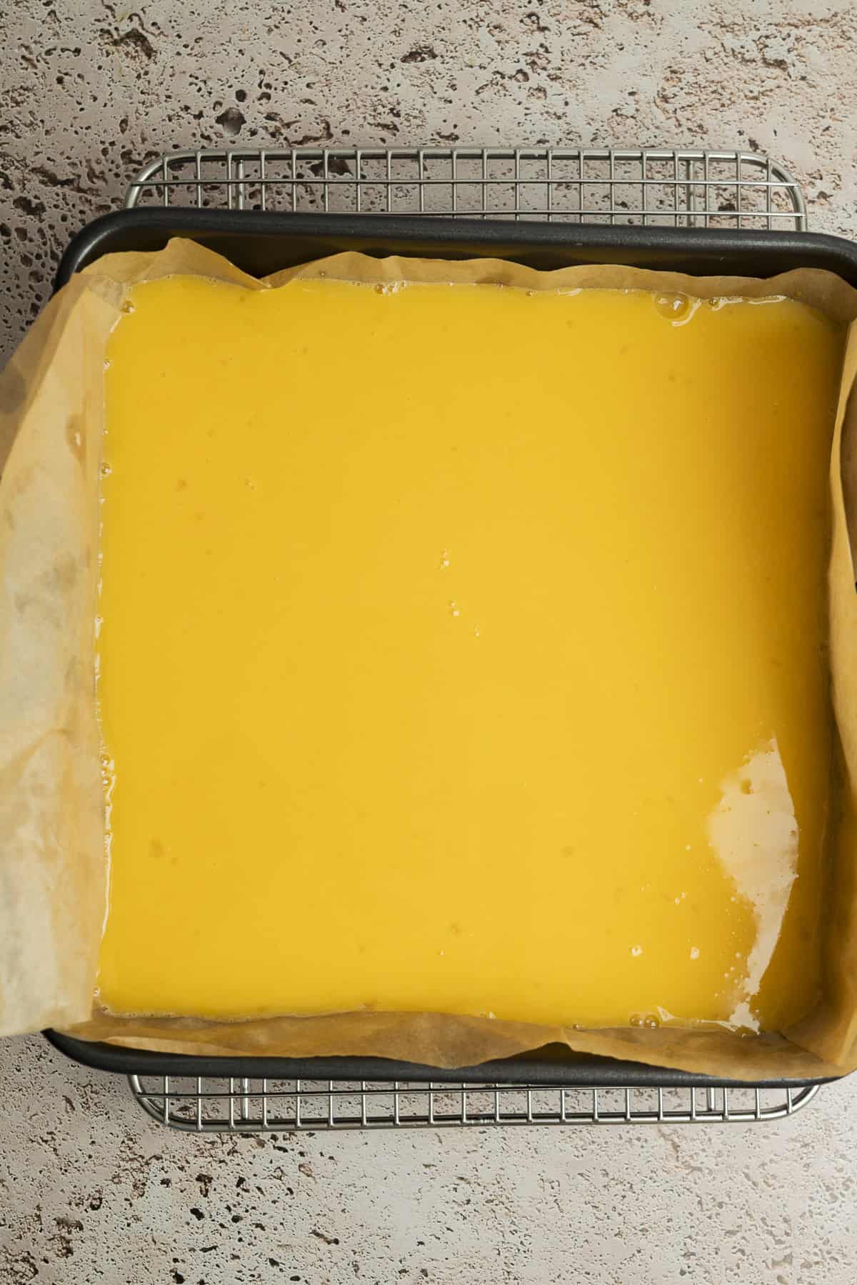 A baking pan lined with parchment paper and filled with a smooth, yellow liquid batter, resting on a cooling rack over a textured countertop.