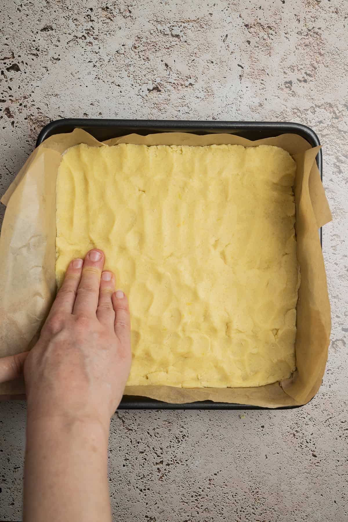 A hand presses down yellow dough evenly into a square baking tin lined with parchment paper on a textured countertop.
