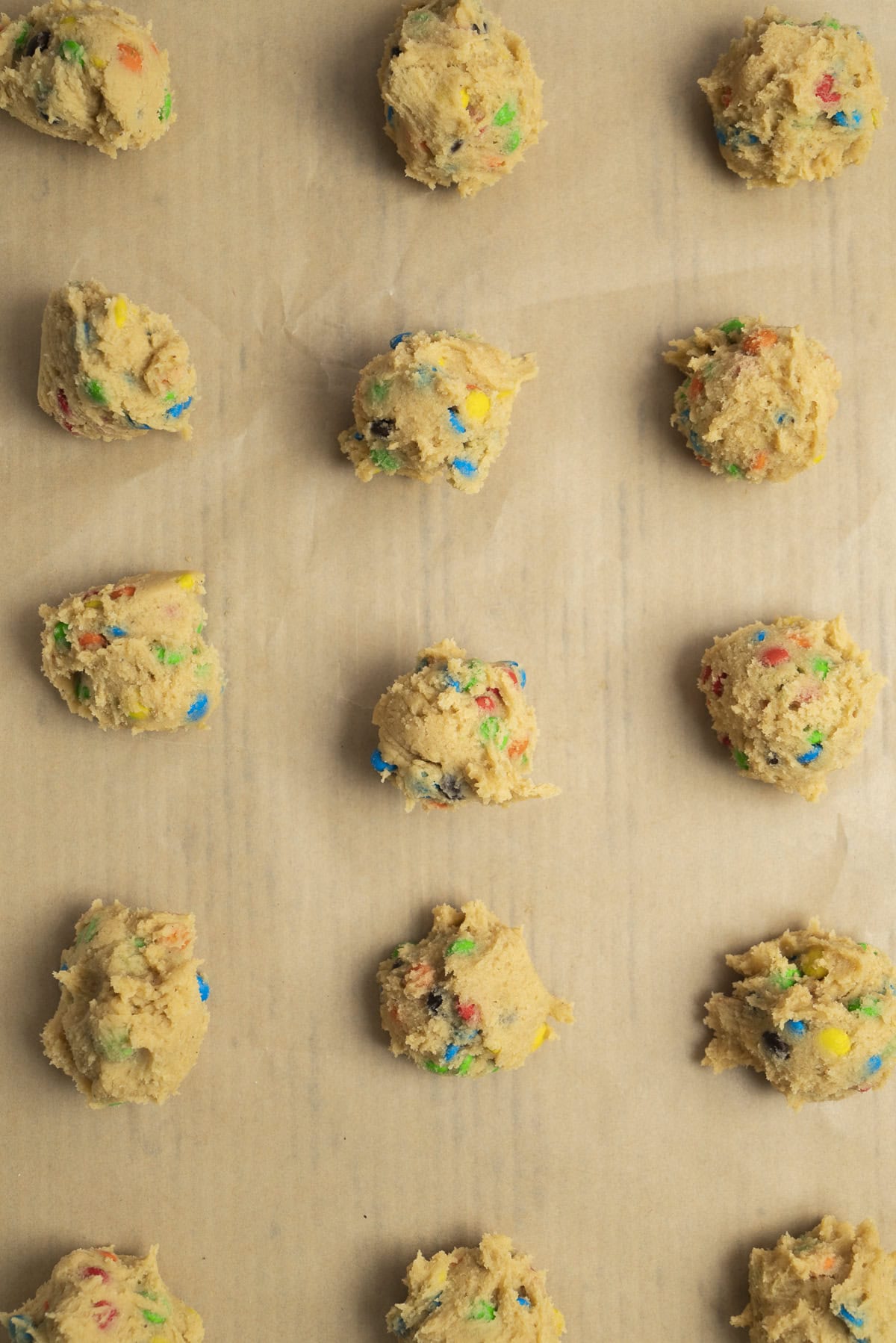 Scoops of cookie dough with colorful candy pieces are evenly spaced on a sheet of parchment paper, ready for baking. The dough is arranged in a neat grid pattern.