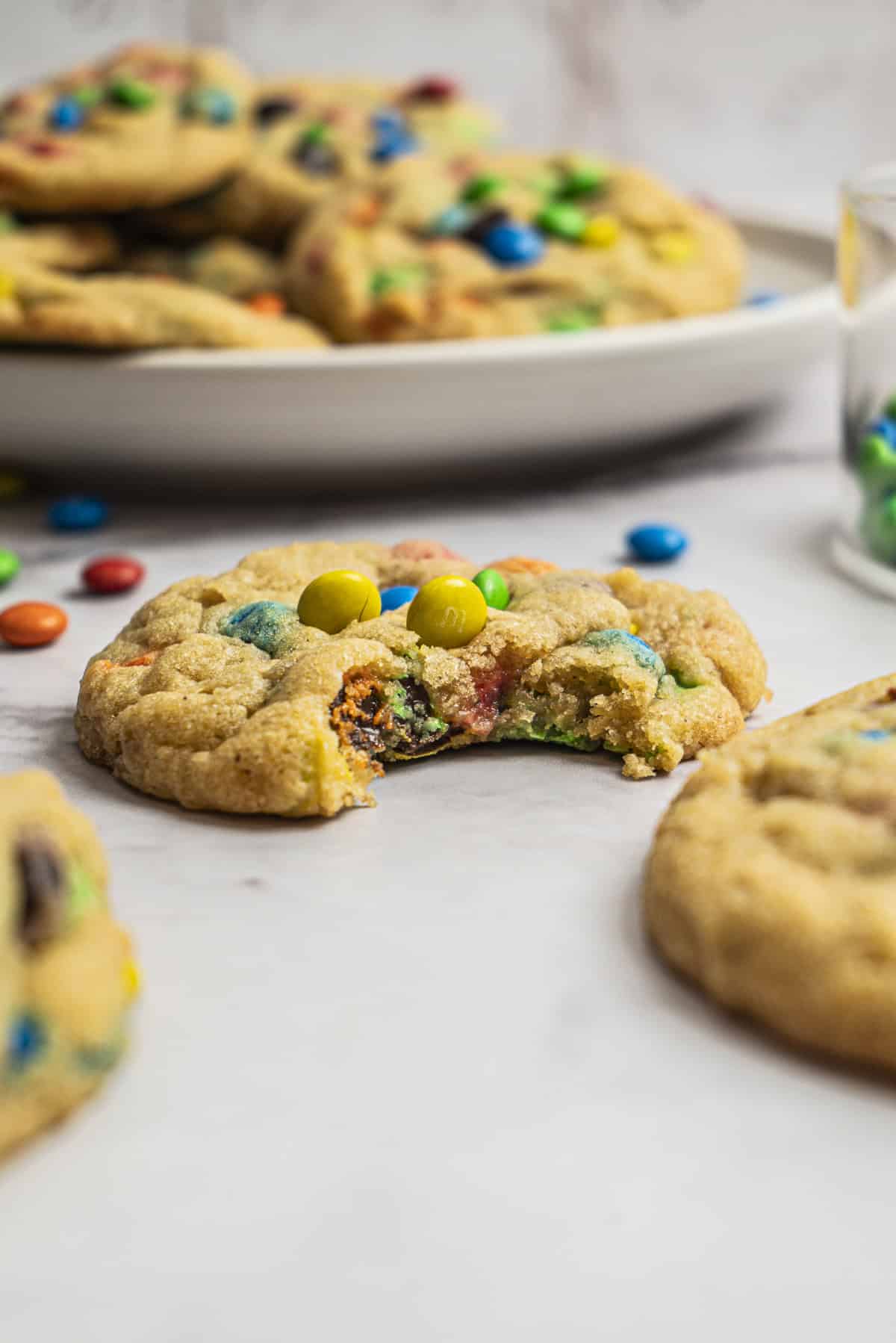 A close-up of a cookie with colorful chocolate candies, partially eaten, sits in the foreground. More whole cookies and scattered candies are visible in the background on a white surface.