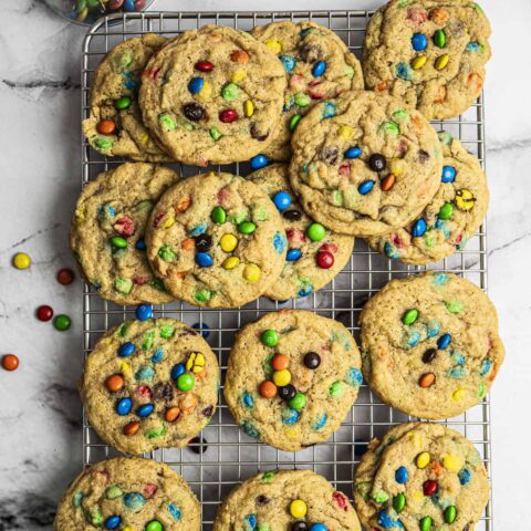 A cooling rack topped with a dozen homemade cookies filled with colorful candy-coated chocolates, with a small glass bowl of more candies nearby on a marble surface.