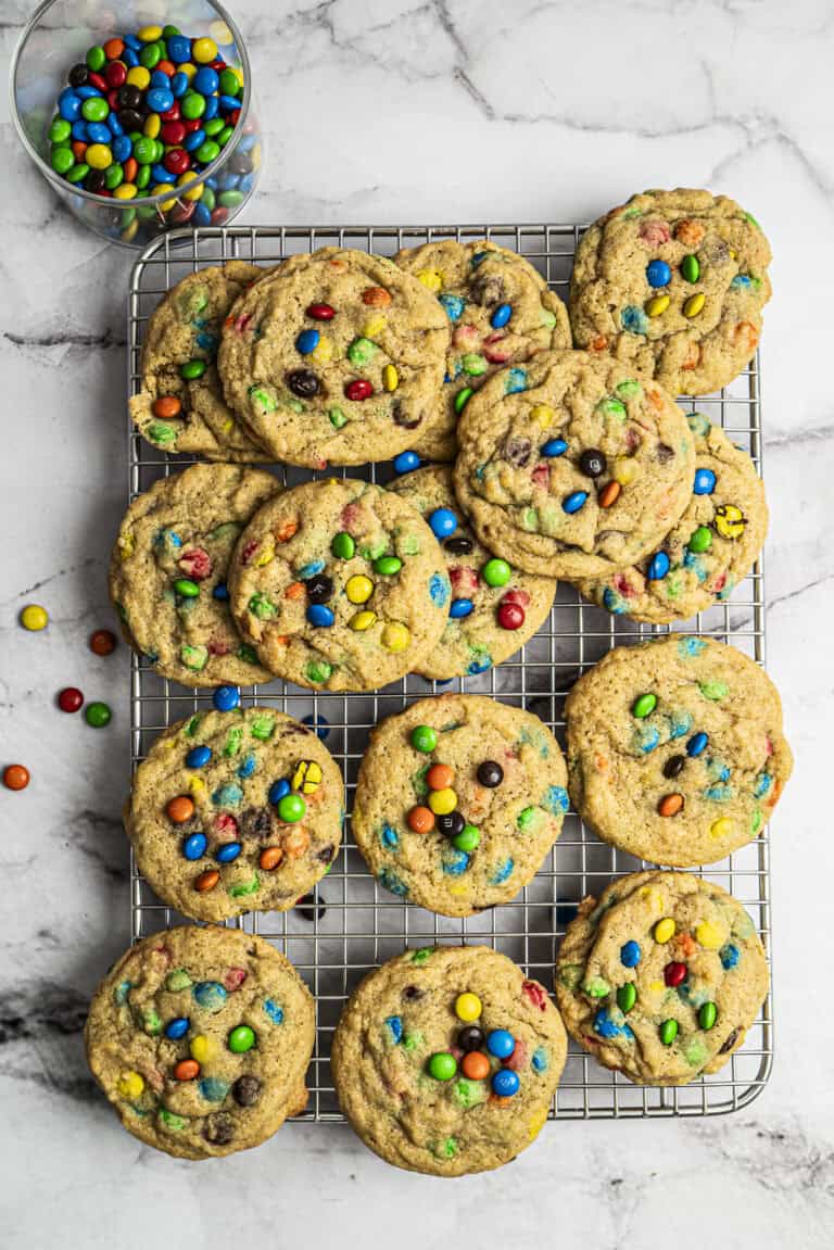 A cooling rack topped with a dozen homemade cookies filled with colorful candy-coated chocolates, with a small glass bowl of more candies nearby on a marble surface.