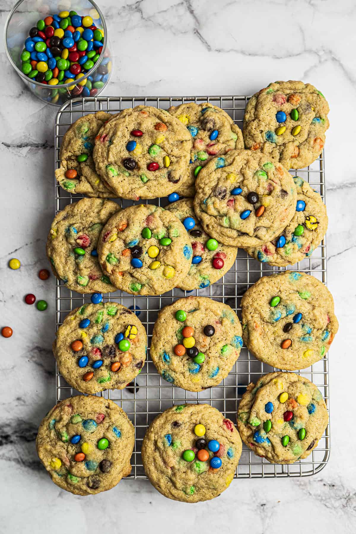 A cooling rack topped with a dozen homemade cookies filled with colorful candy-coated chocolates, with a small glass bowl of more candies nearby on a marble surface.