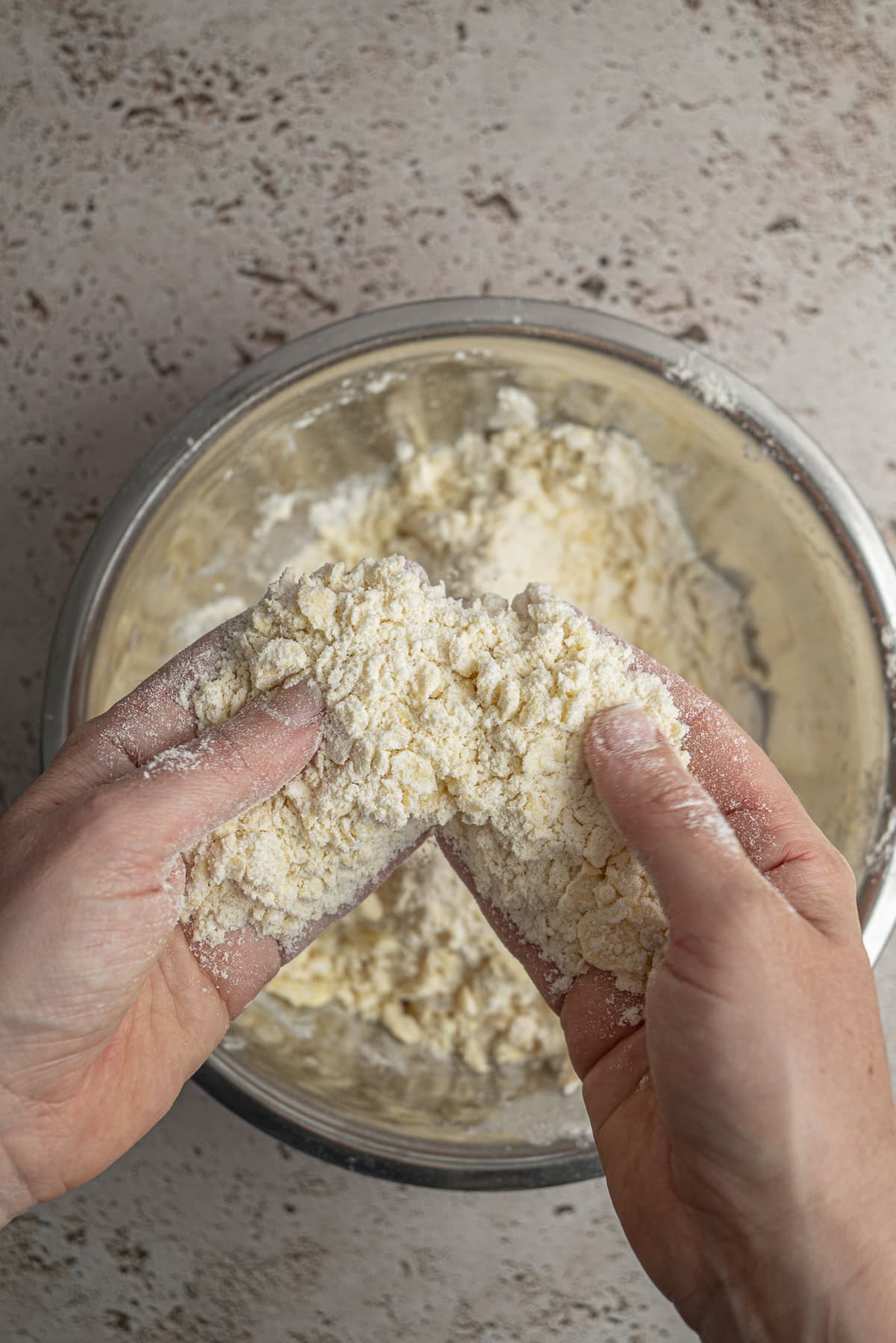 Two hands rub flour and butter together over a metal mixing bowl, creating a crumbly dough mixture for delicious Blackberry Hand Pies on a speckled countertop.