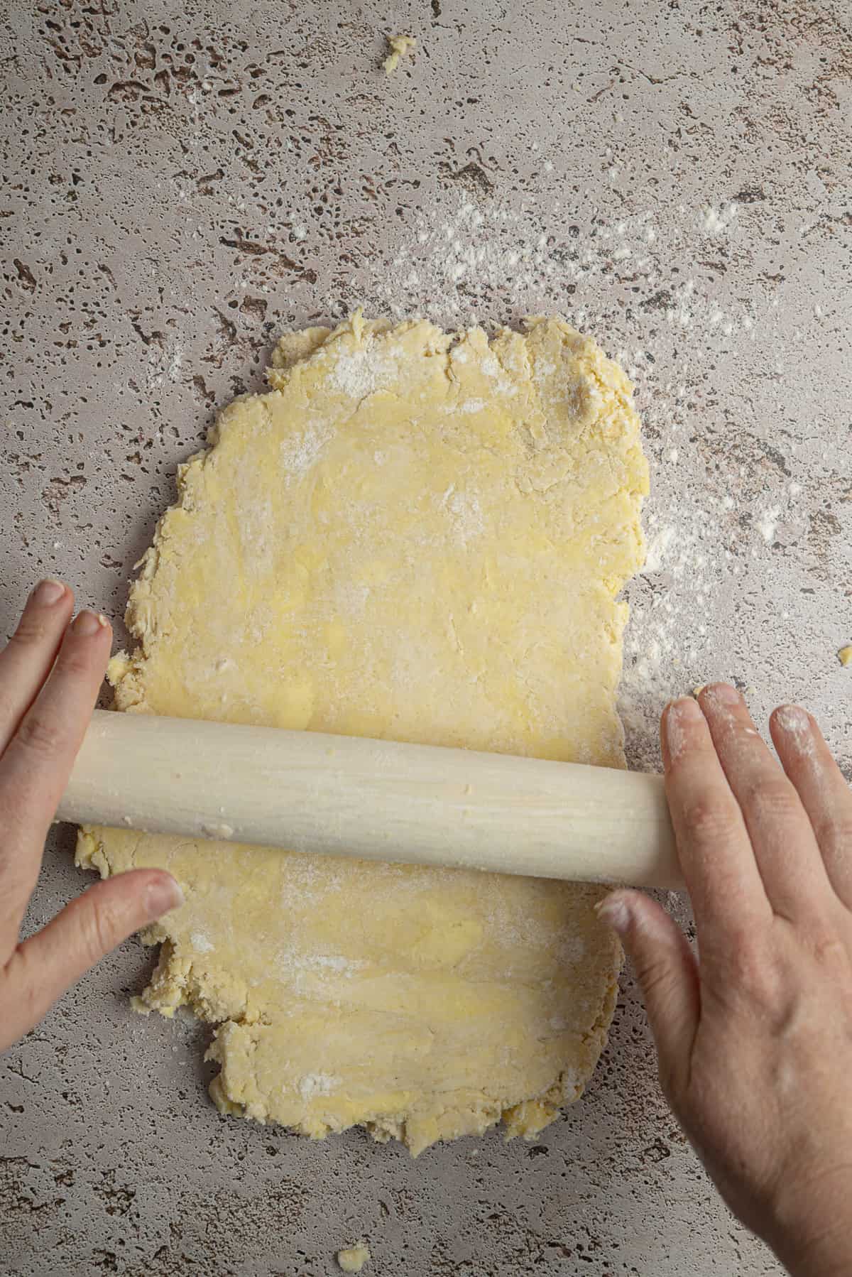 A person rolls out a sheet of yellow dough with a wooden rolling pin on a floured, textured surface, preparing it to make delicious Blackberry Hand Pies.