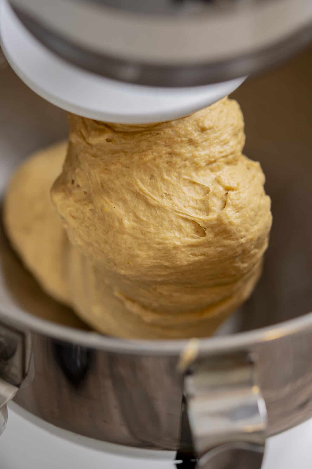 A close-up of bread dough being mixed in a stand mixer with a dough hook attachment inside a metal mixing bowl.