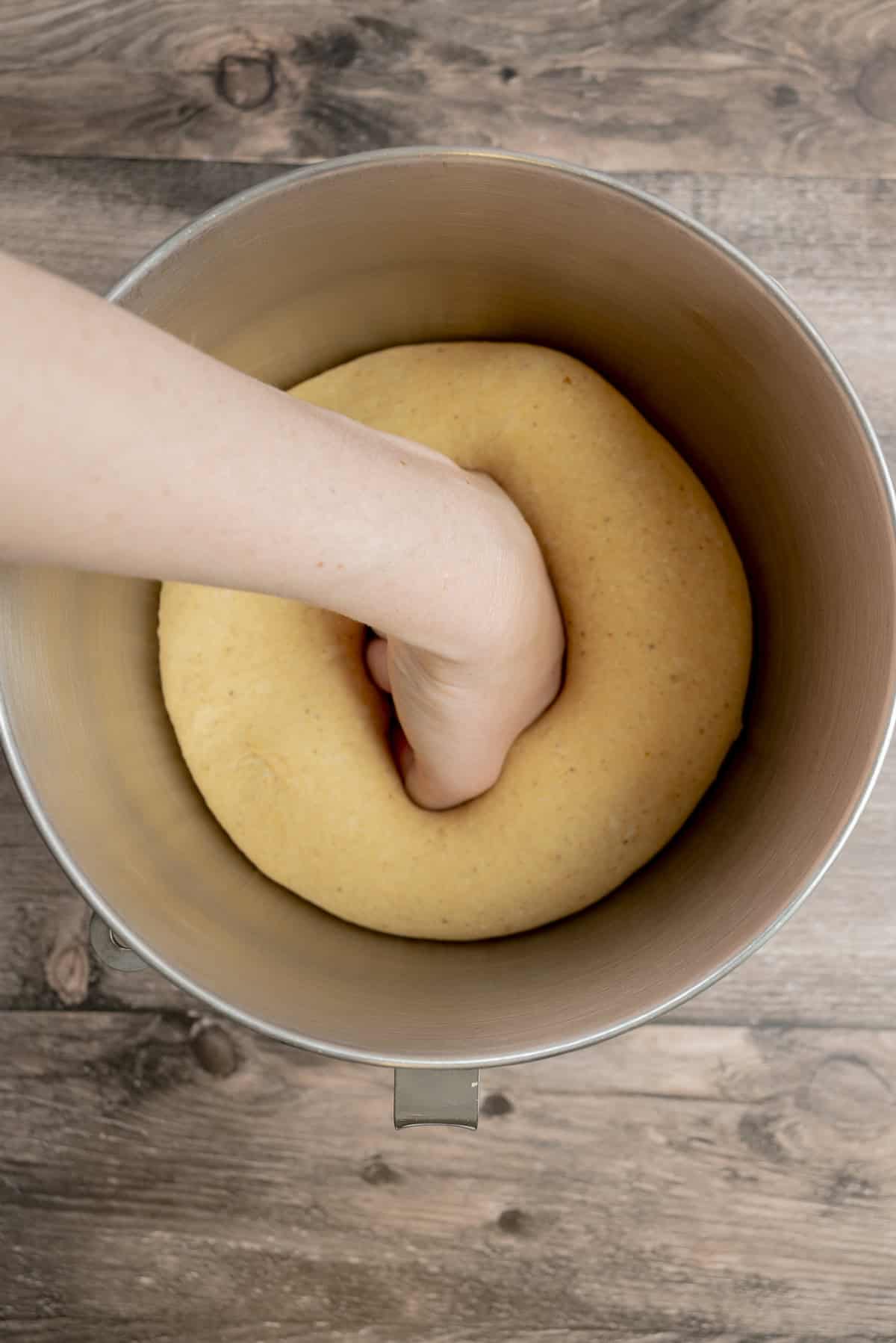 A hand presses down into risen dough inside a large metal mixing bowl on a wooden surface.