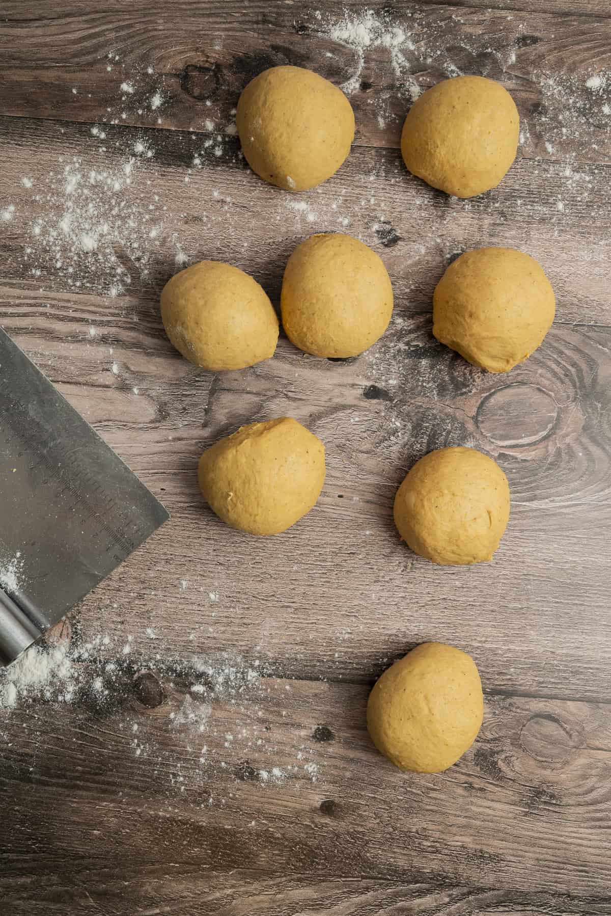 Eight round balls of dough are arranged on a floured wooden surface with a metal dough scraper on the left side of the image.