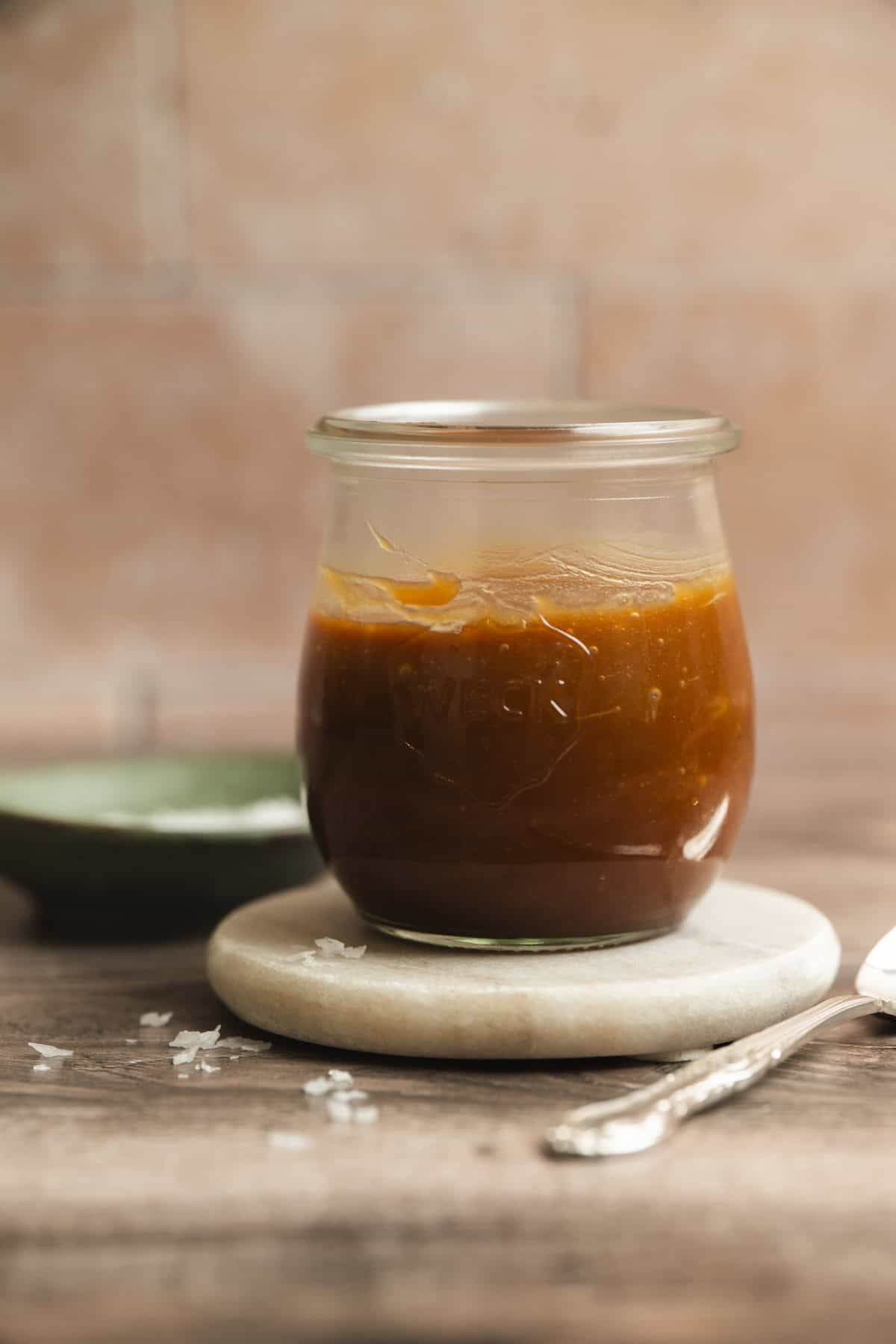 A glass jar filled with a small batch caramel sauce sits on a round marble coaster, with a fork and some sea salt flakes nearby on a wooden table. The background is softly blurred with warm, earthy tones.