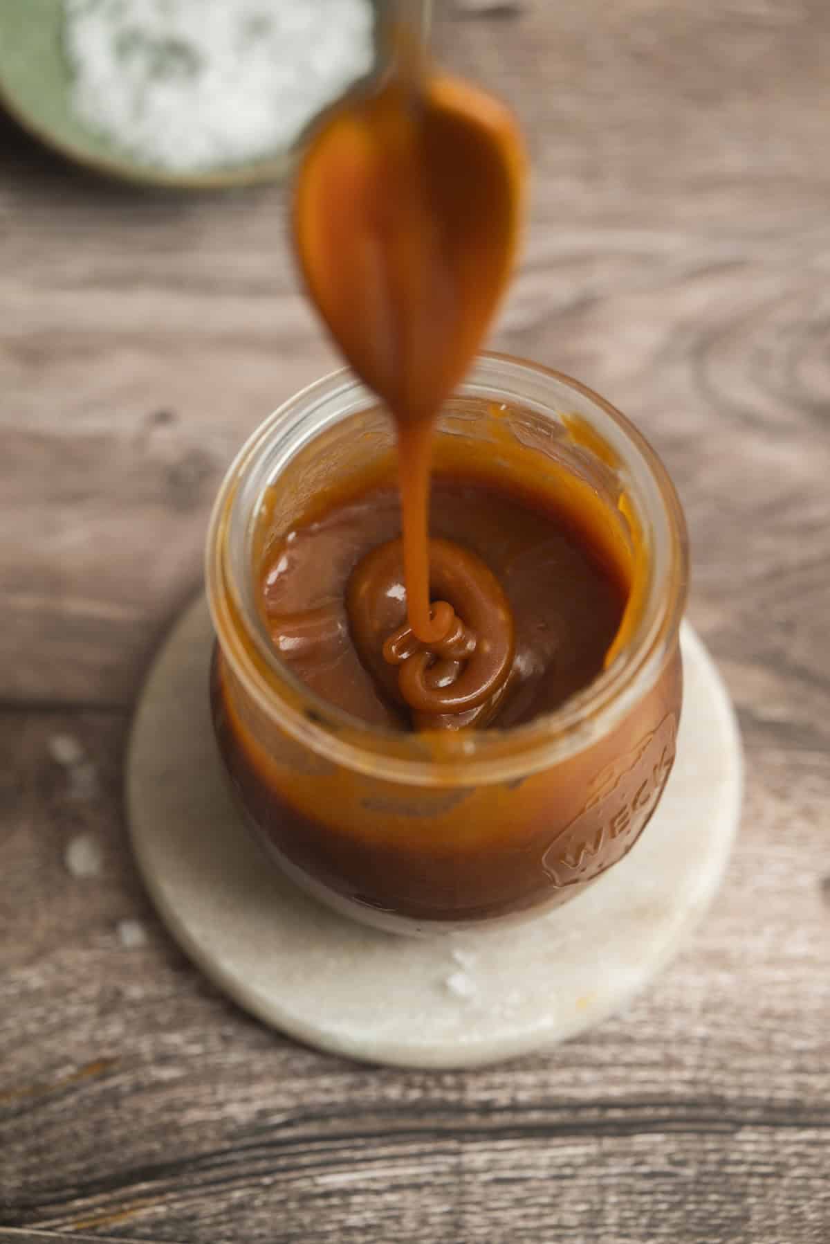 A spoon drizzles thick small batch salted caramel sauce into a glass jar filled with caramel, set on a round marble coaster atop a wooden surface.