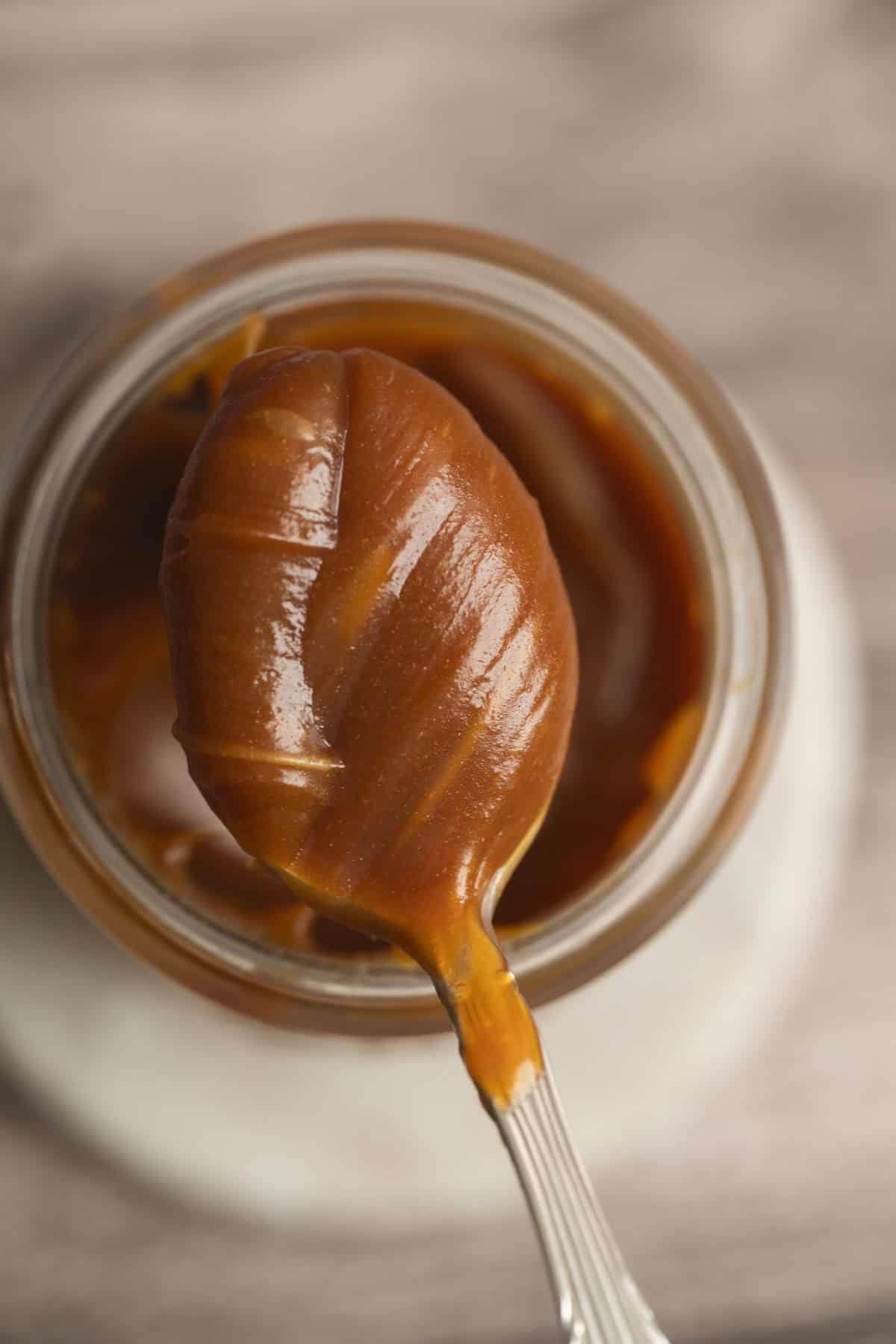 A close-up of a spoon covered in thick, rich salted caramel sauce held above an open glass jar filled with more caramel sauce.