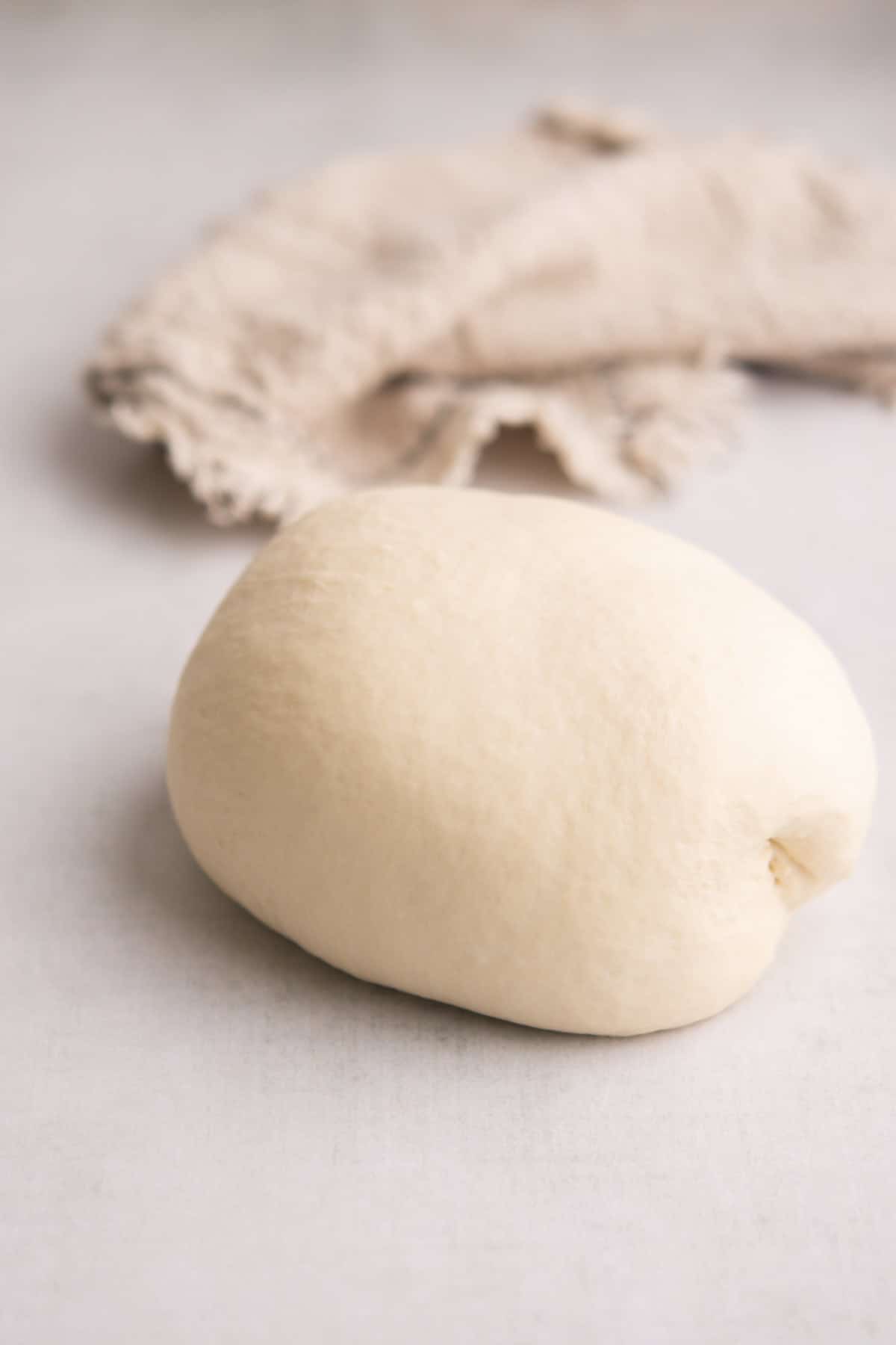 A smooth ball of dough sits on a light surface with a beige, textured cloth in the background.