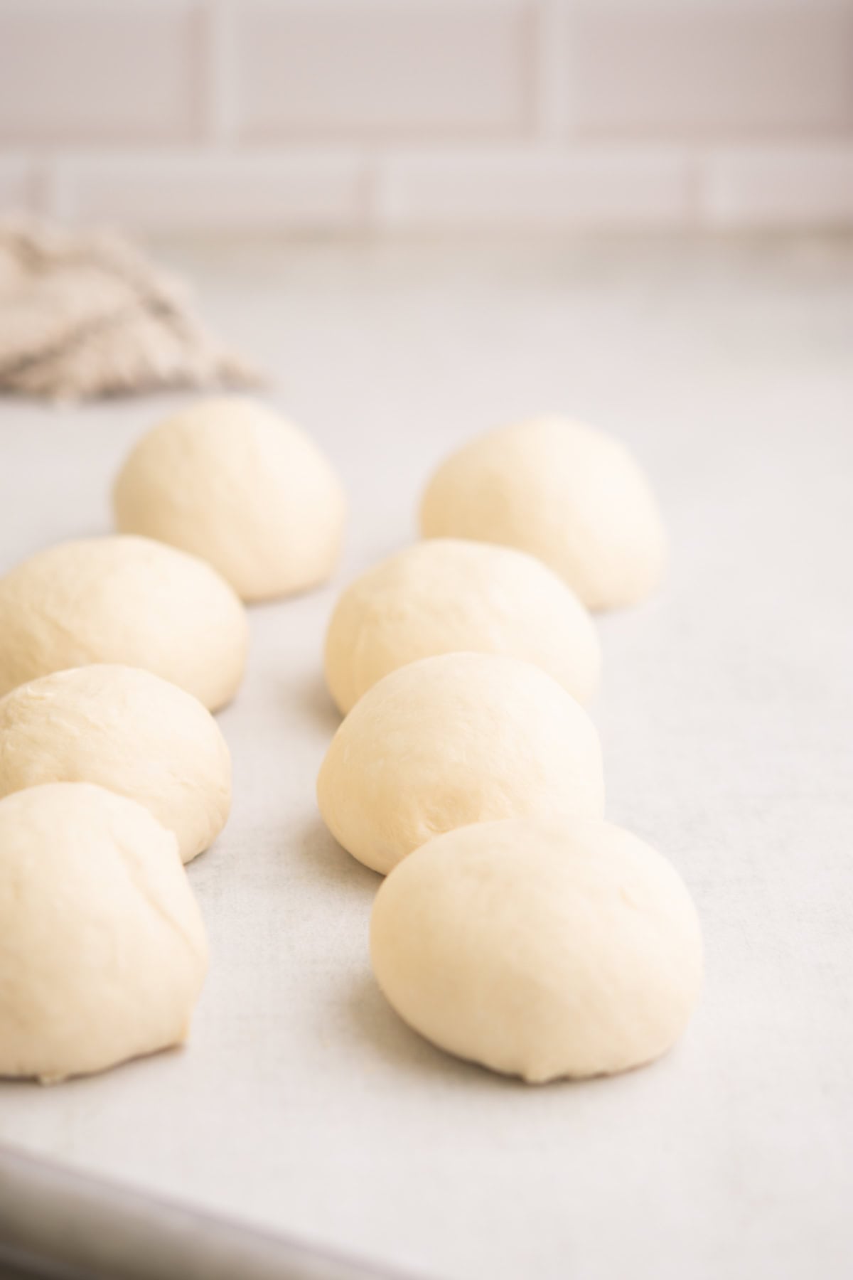 Eight smooth, round balls of dough are arranged on a light-colored countertop with a soft, neutral background and a blurred cloth in the corner.