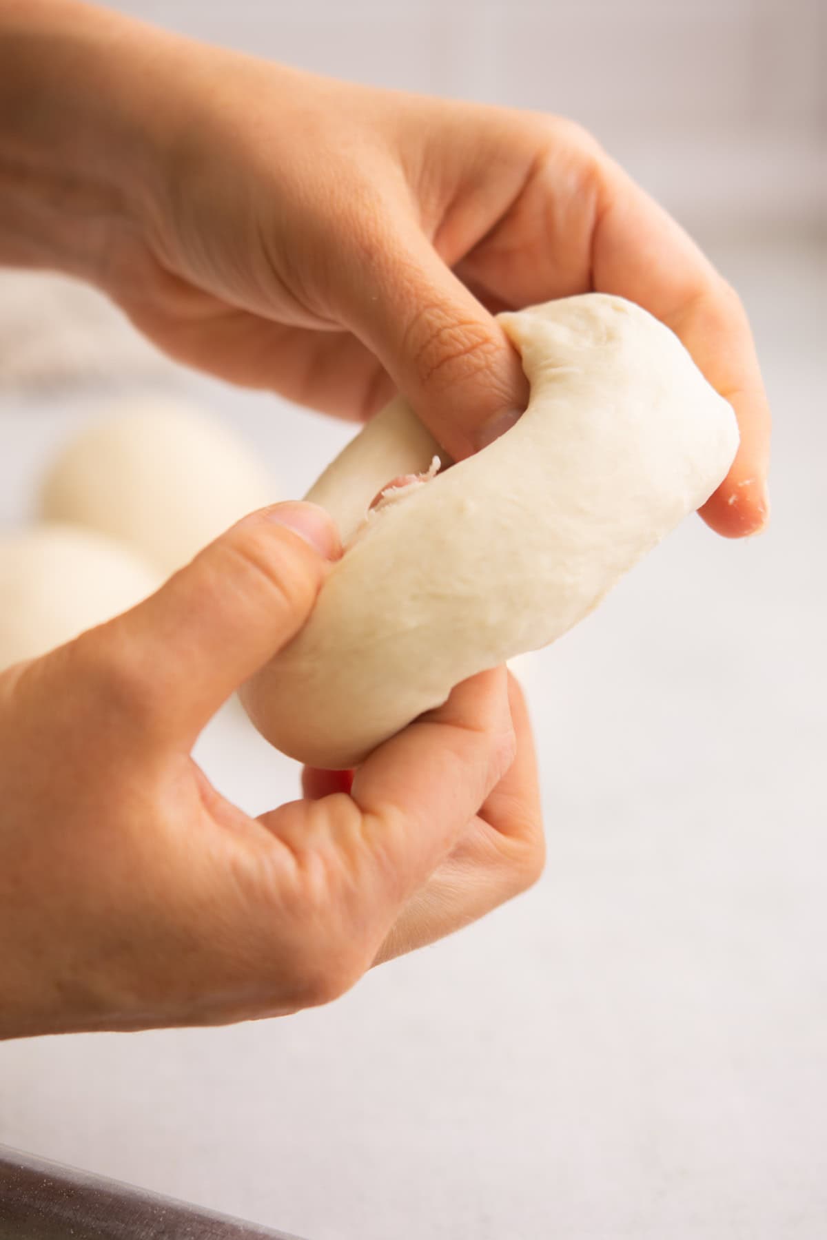 Close-up of hands shaping a piece of dough into a ring on a light surface, with other dough balls blurred in the background.