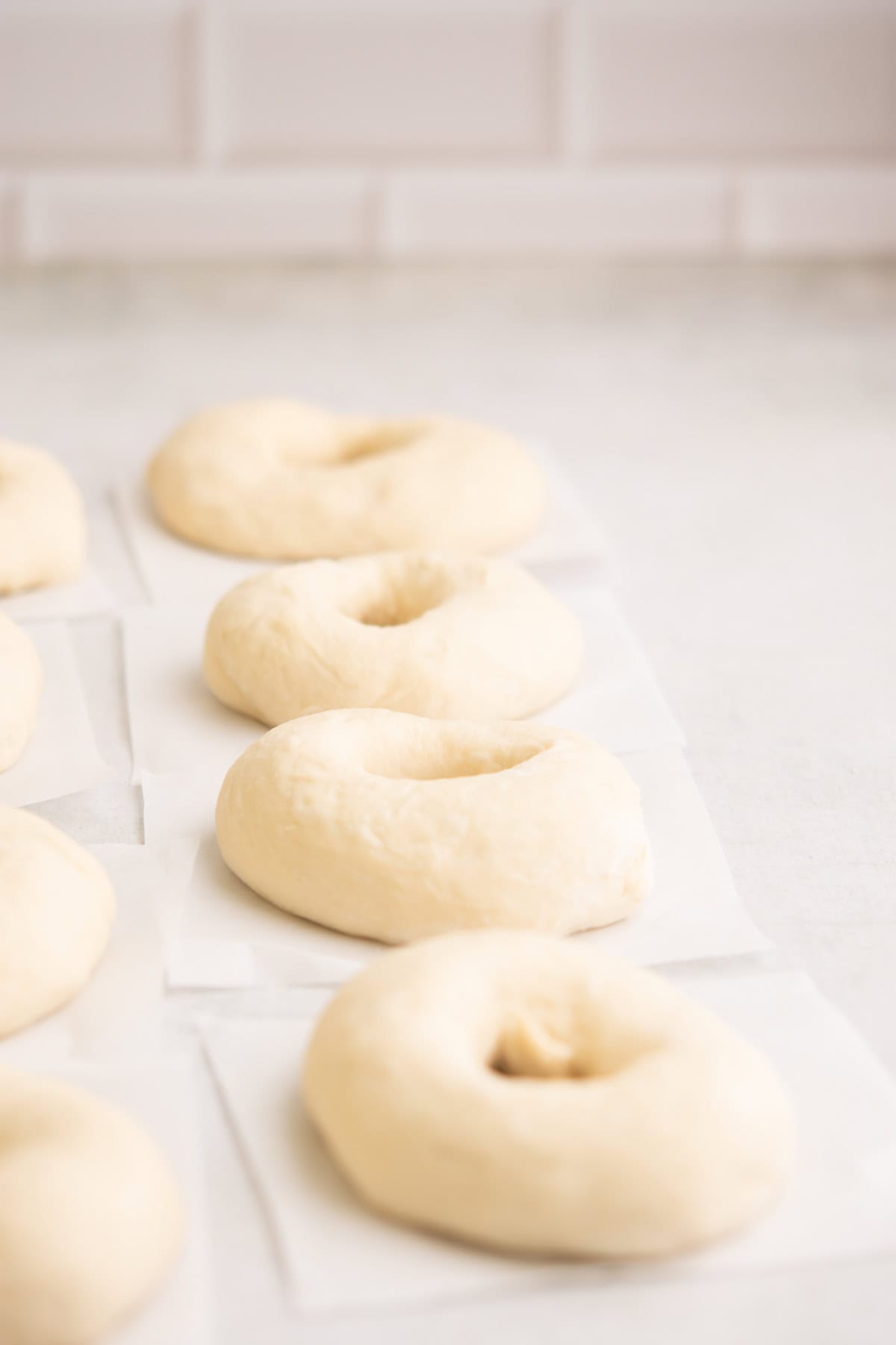 Several unbaked bagels made from pale dough rest on small squares of parchment paper on a light-colored countertop, ready for baking. The background is softly blurred.