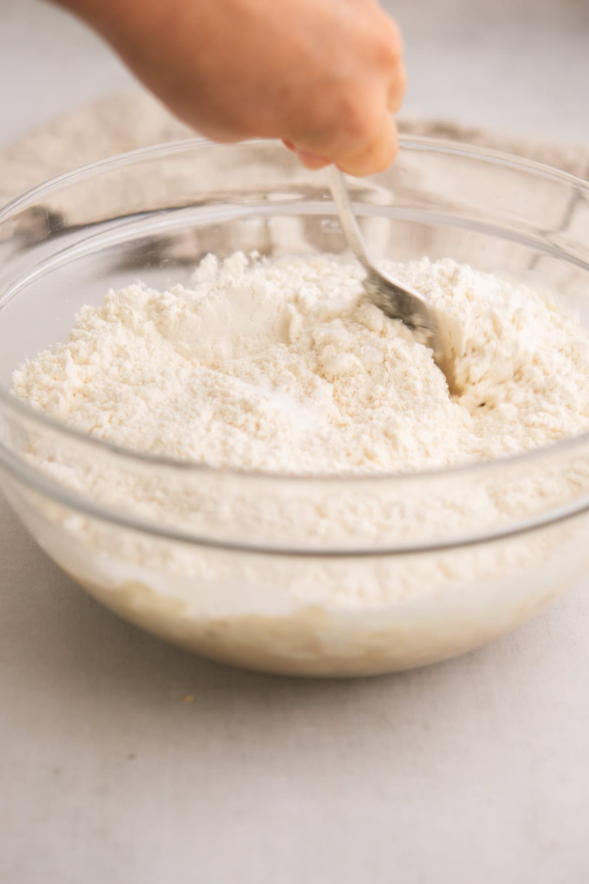 A hand stirring a clear glass bowl filled with flour using a metal spoon, on a light-colored surface.