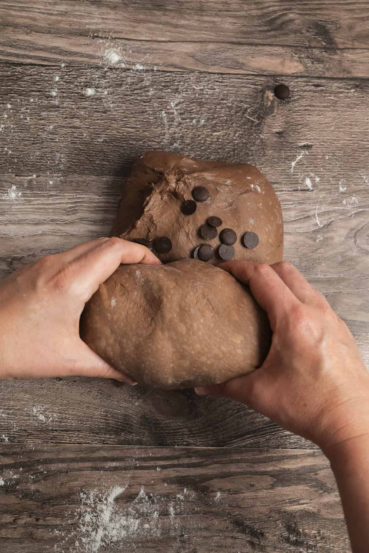 Two hands kneading chocolate dough with chocolate chips on a wooden surface dusted with flour.