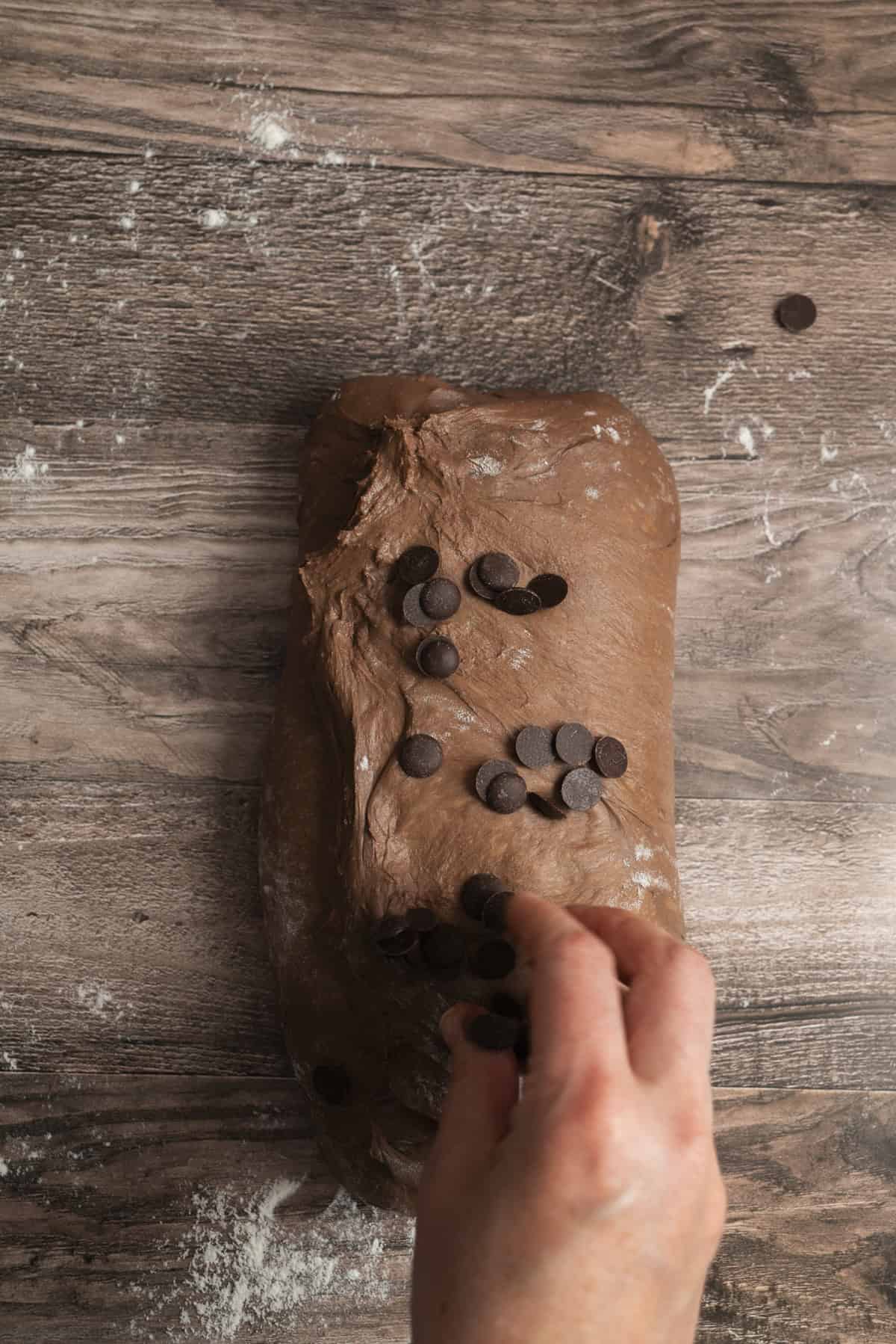 A hand adds chocolate chips to a loaf of chocolate dough on a floured wooden surface, preparing it for baking.