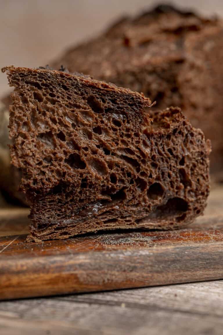 A close-up of a slice of dark chocolate sourdough bread with a porous, airy texture, resting on a wooden cutting board. Another piece of bread is visible in the blurred background.