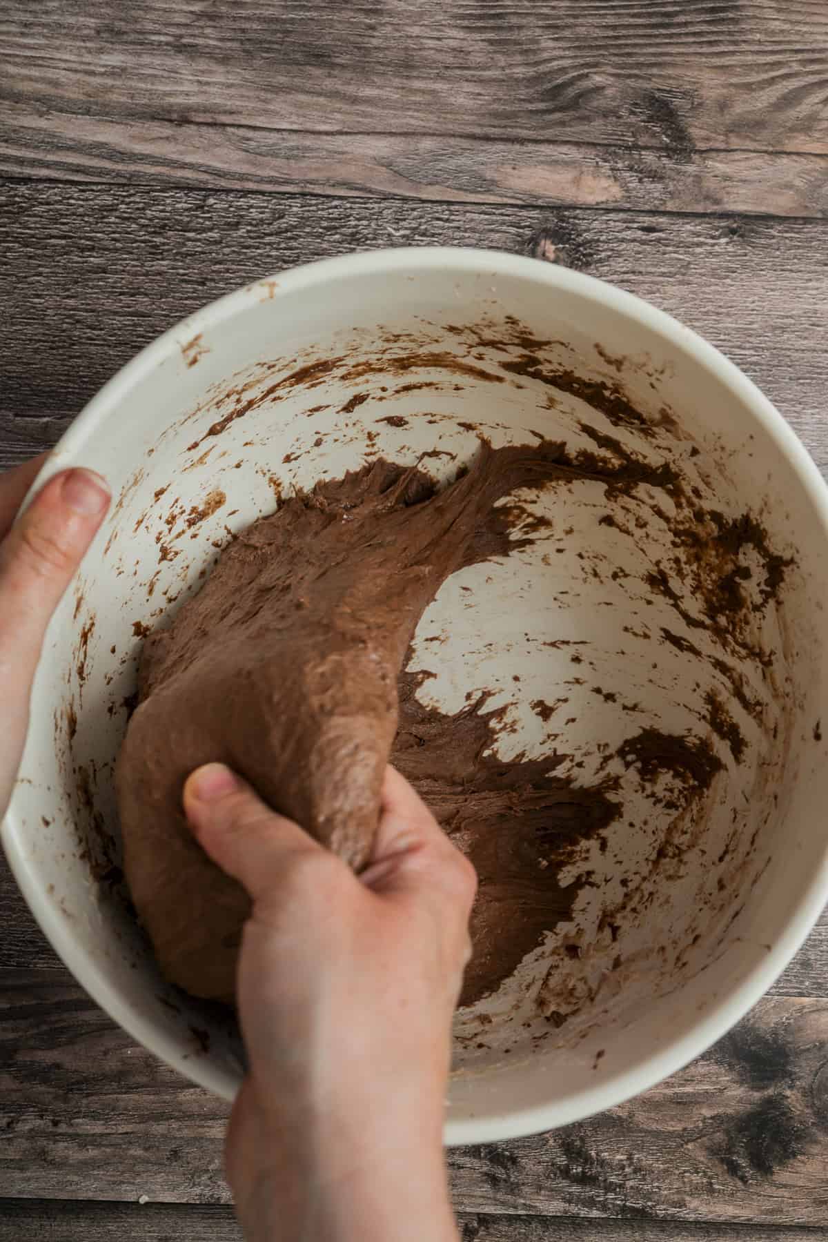 Two hands knead chocolate dough inside a large white bowl, with dough residue on the sides. The bowl rests on a wooden surface.