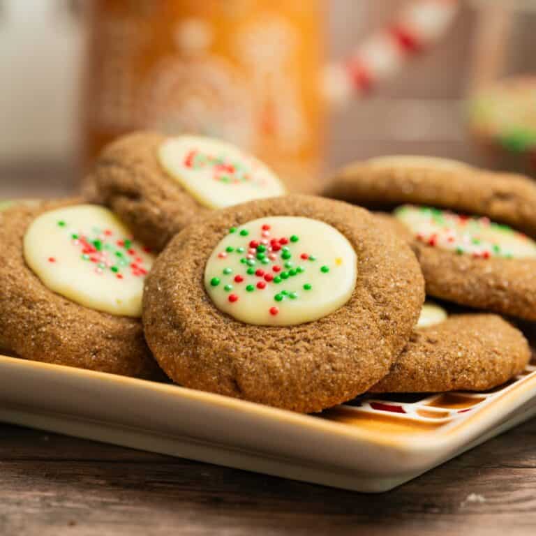 A plate of gingerbread thumbrints with white chocolate ganache and red and green sprinkles, arranged on a square dish, with a blurred festive background.