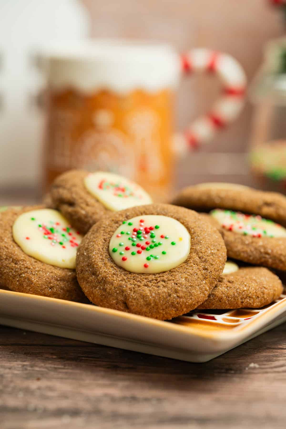 A plate of ginger cookies topped with white icing and red and green sprinkles, with a festive mug and holiday decorations blurred in the background.