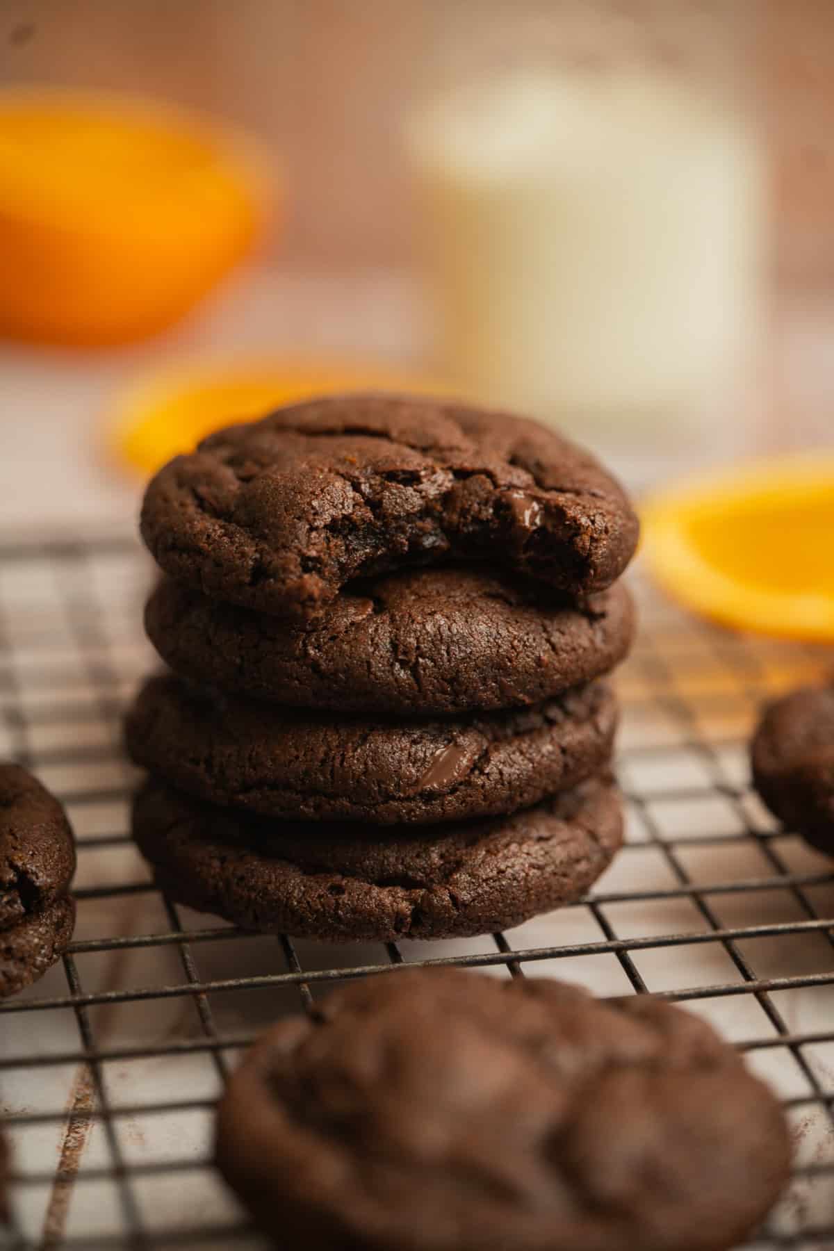 A stack of five chocolate cookies, with the top cookie missing a bite, sits on a cooling rack. Blurred in the background are an orange slice and a glass of milk.