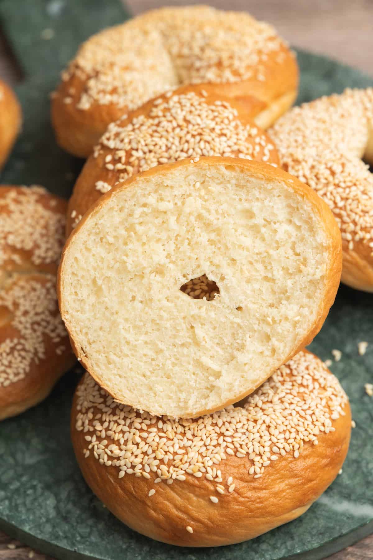 A sliced sesame bagel with a soft, airy crumb is stacked on top of whole sesame bagels on a green plate. The bagels are golden-brown and generously topped with sesame seeds.