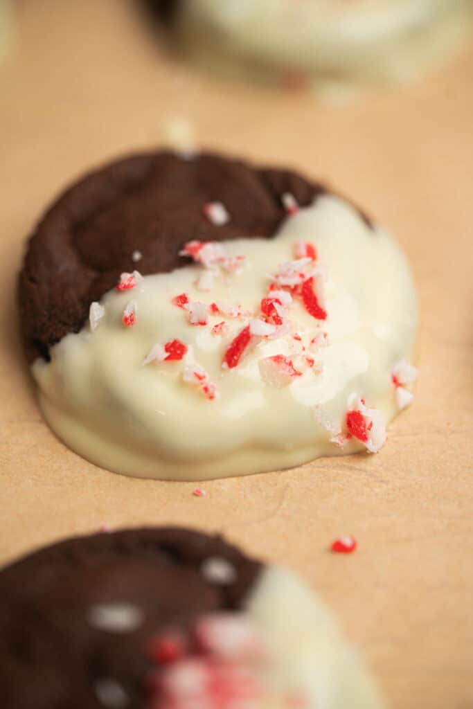 A chocolate cookie half-dipped in white chocolate and sprinkled with crushed peppermint candy, resting on a sheet of parchment paper.
