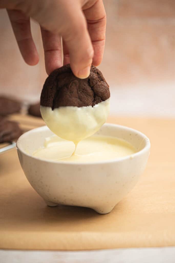 A hand dips a chocolate cookie halfway into a bowl of white melted chocolate, with some chocolate dripping off. The bowl sits on a tan surface, and more cookies are visible in the background.