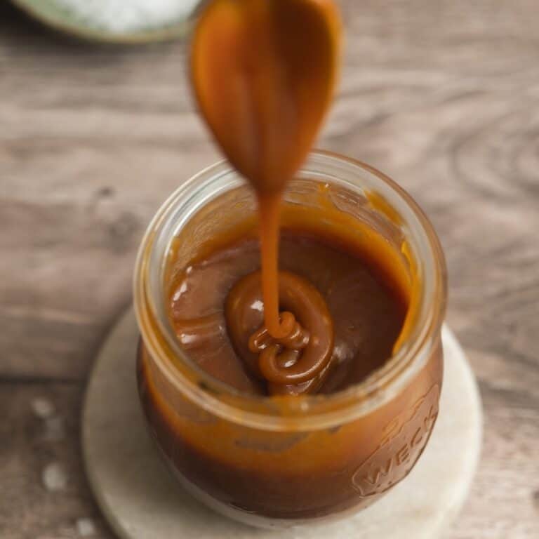 A spoon drizzles thick caramel sauce into a glass jar filled with caramel, set on a round coaster atop a wooden surface.