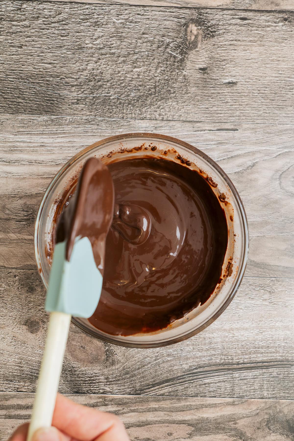 A hand holds a spatula coated in melted chocolate above a glass bowl filled with smooth, glossy melted chocolate on a wooden surface.