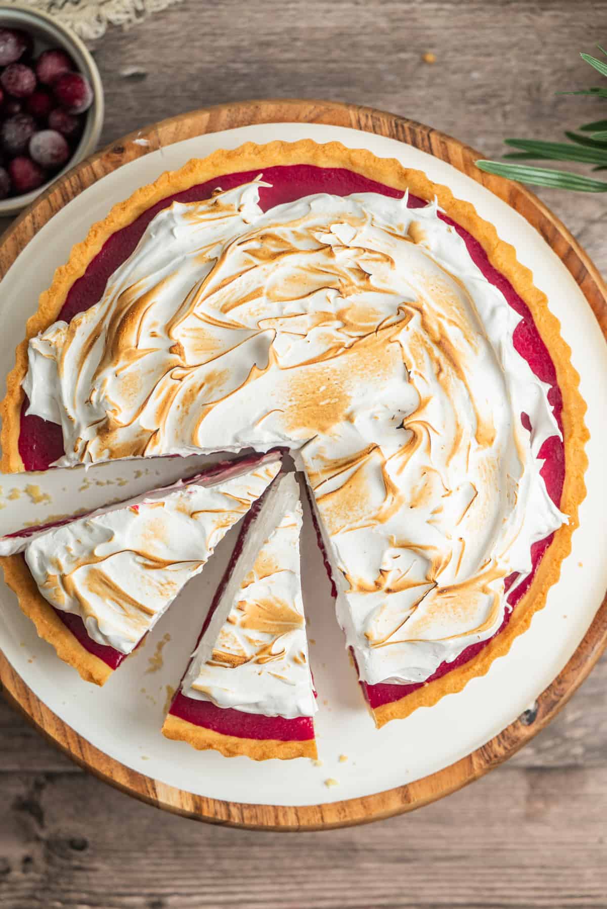 A cranberry tart topped with toasted swirls of meringue sits on a wooden platter. Two slices have been cut, showing the vibrant red filling beneath the meringue. A bowl of cranberries is nearby.