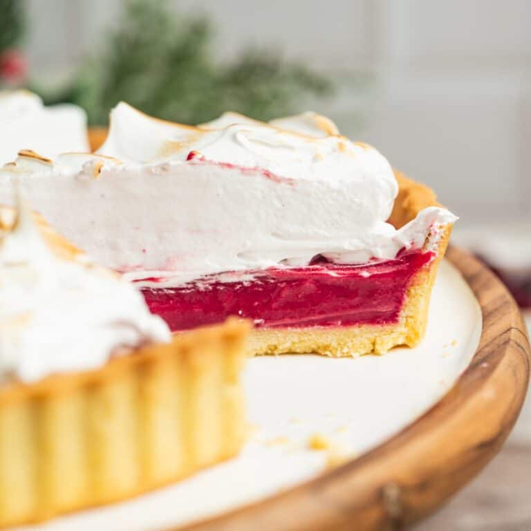 A close-up of a tart with a thick, bright red cranberry filling and a golden crust, topped with a layer of fluffy white meringue. A slice has been removed, showing the vibrant filling and texture.
