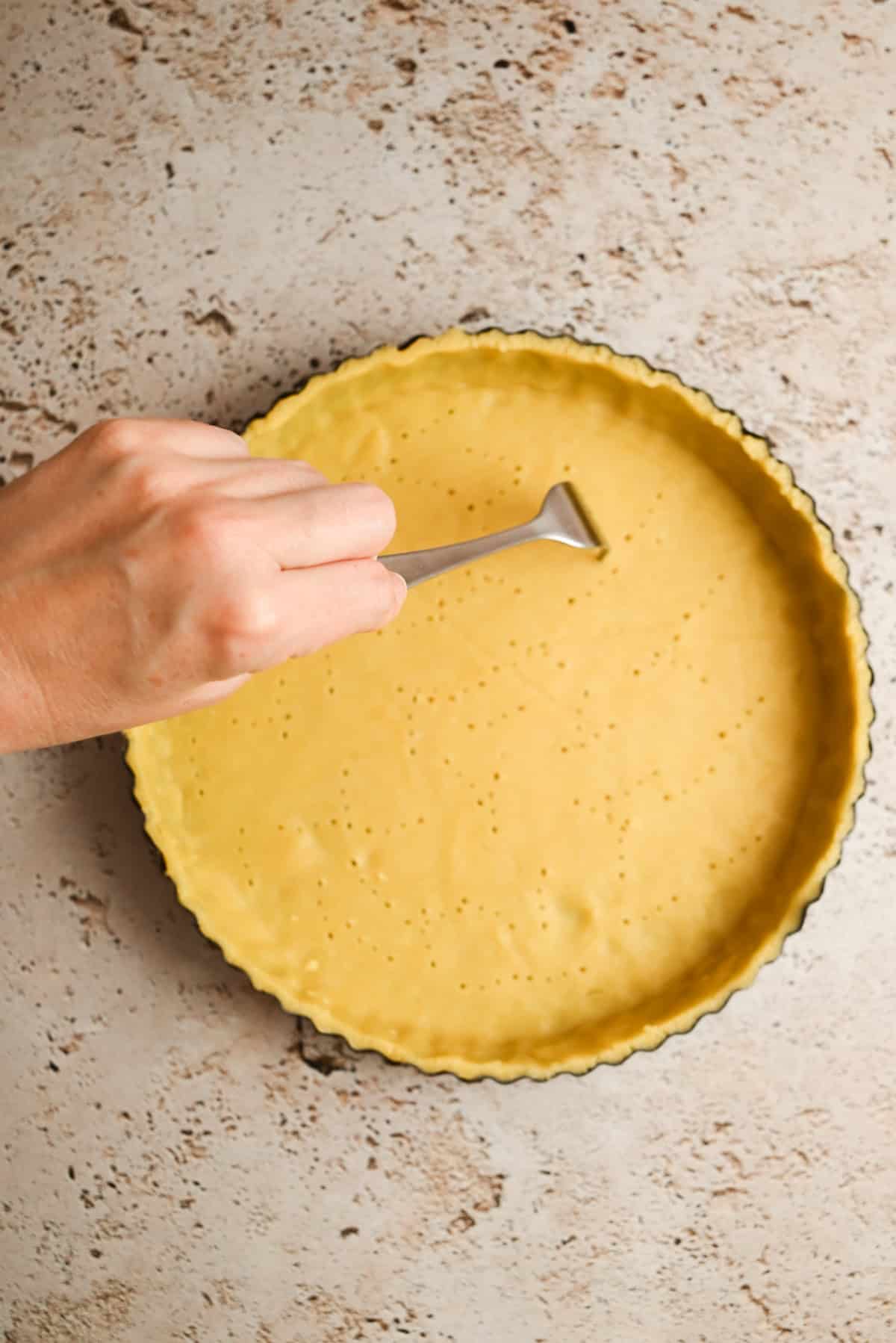 A hand uses a fork to prick holes in an unbaked pie crust in a round tart pan on a light, textured surface.