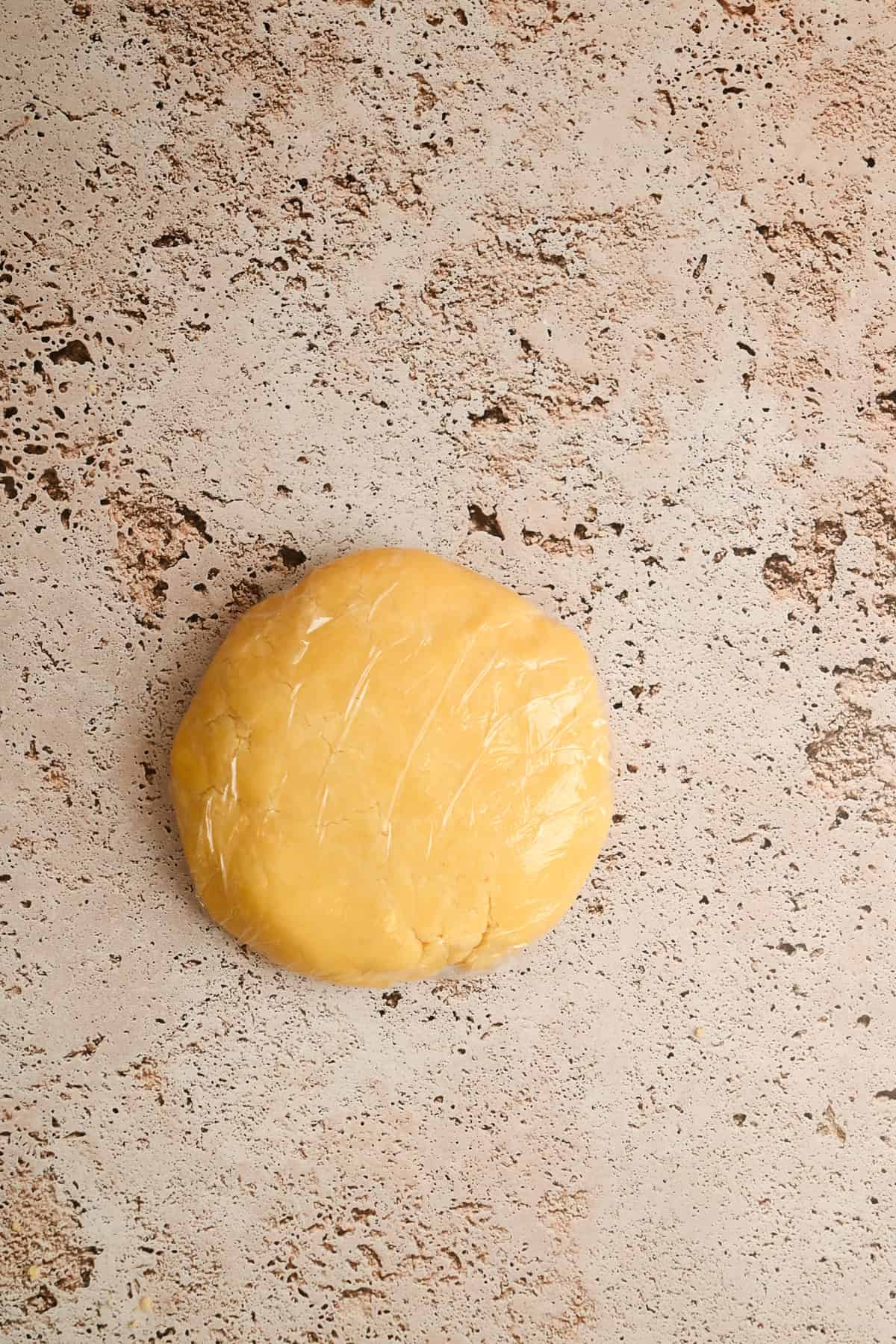 A round disk of dough wrapped in plastic wrap rests on a light, textured surface.