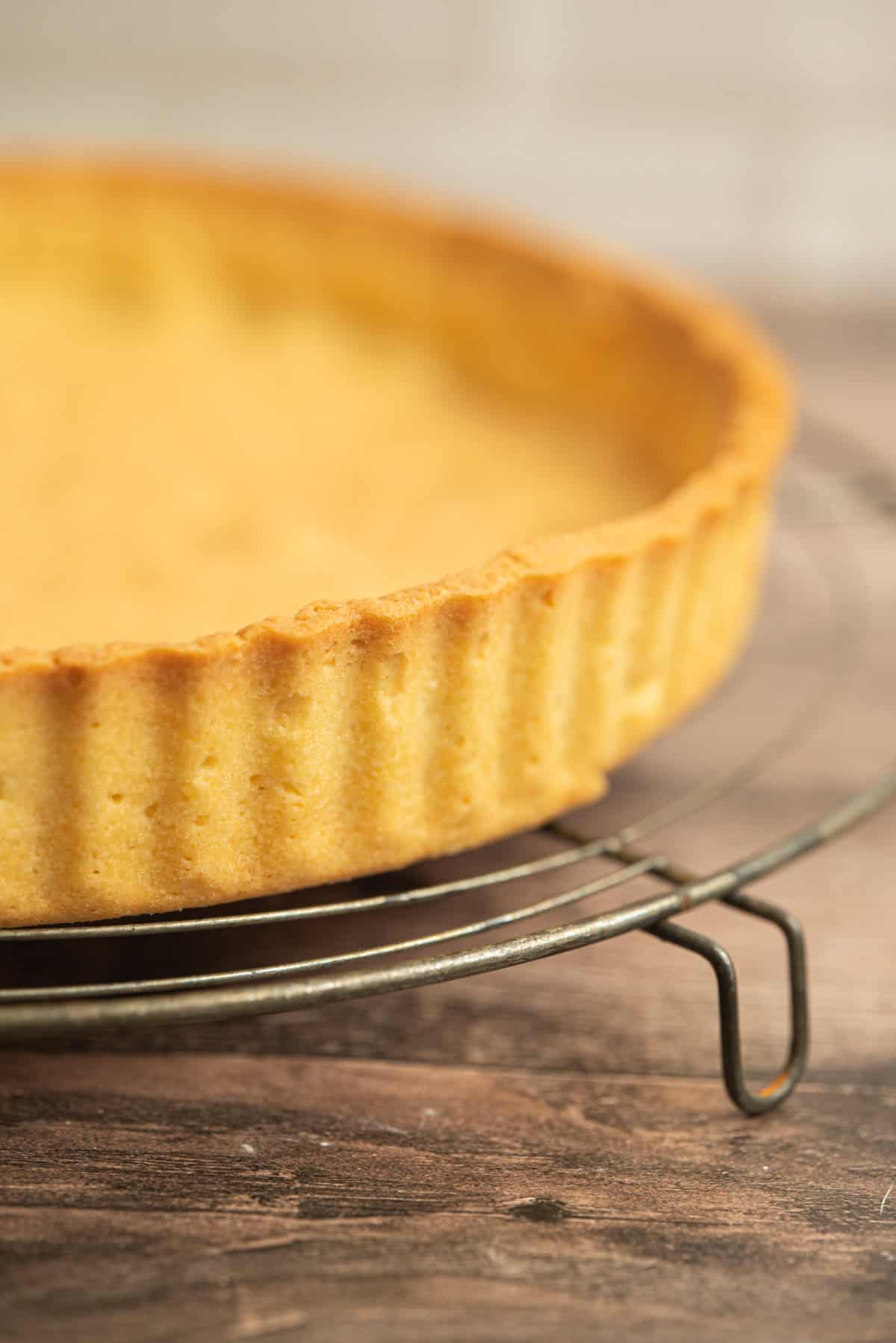A close-up of a baked pate sablee tart crust on a metal cooling rack, with a golden-brown edge and a textured, fluted rim, sitting on a wooden surface.